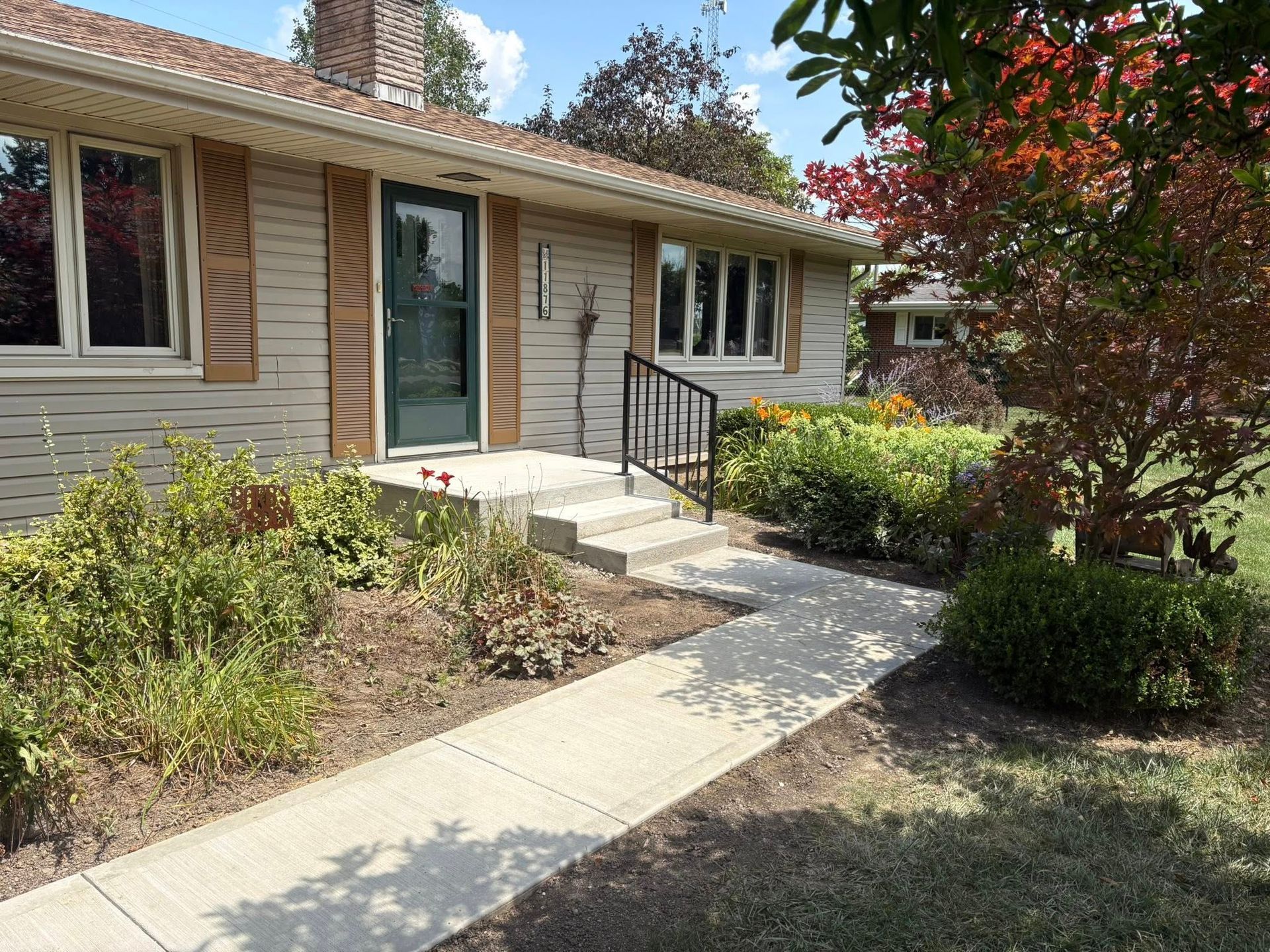 A tan single-story house with brown shutters, a green front door, and a concrete walkway leading to the entrance.