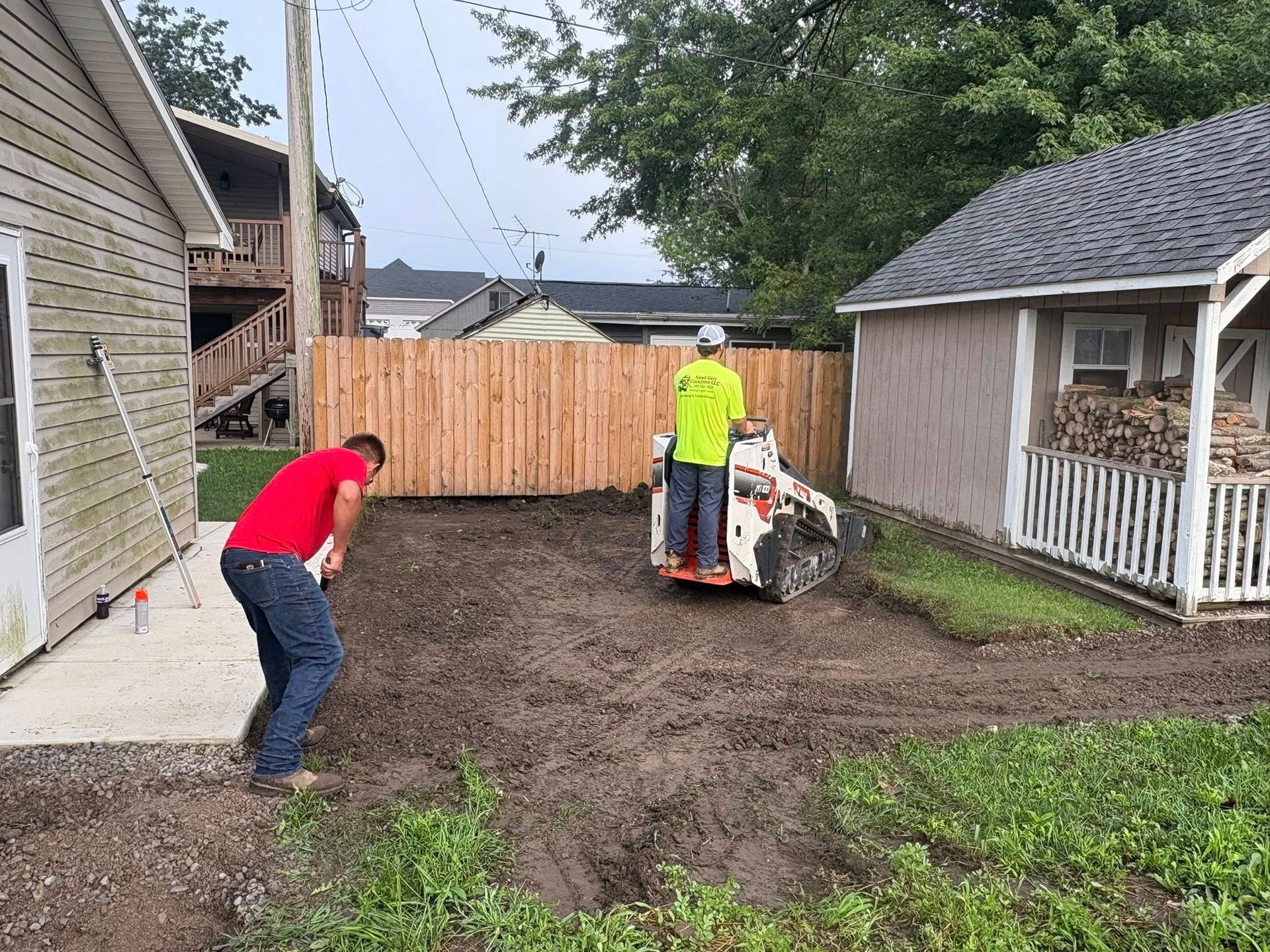 Two people work on a muddy residential yard, one operating a small skid-steer loader and the other holding a tool.