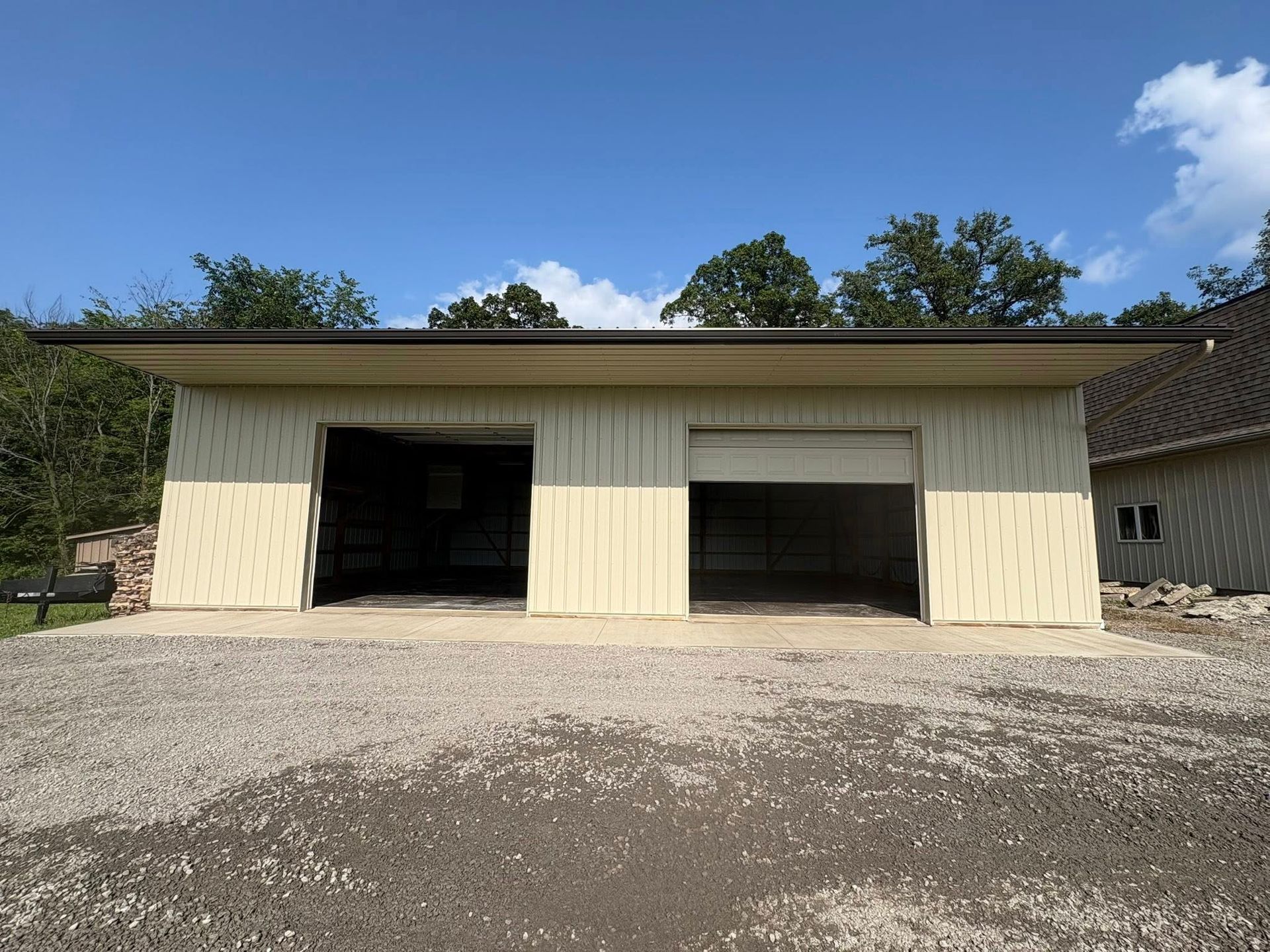A light-tan, two-bay metal garage with open doors sitting on a gravel lot under a clear blue sky.