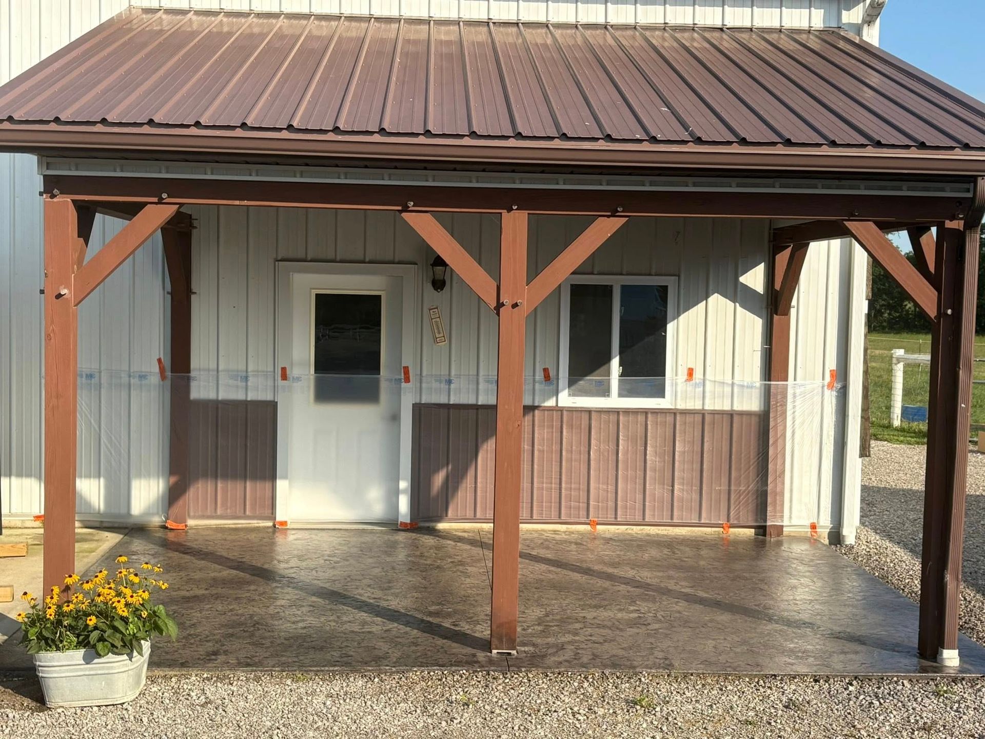 A covered porch with a dark-patterned concrete patio, white door, window, and brown support posts in front of a building.