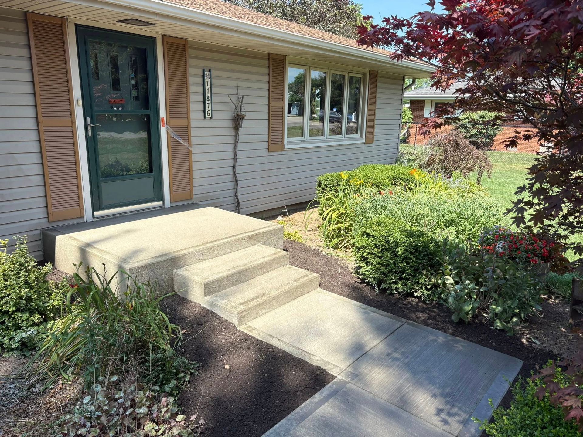 A light gray house exterior with a concrete porch, two stairs, a dark green storm door, and landscaped front yard bushes.