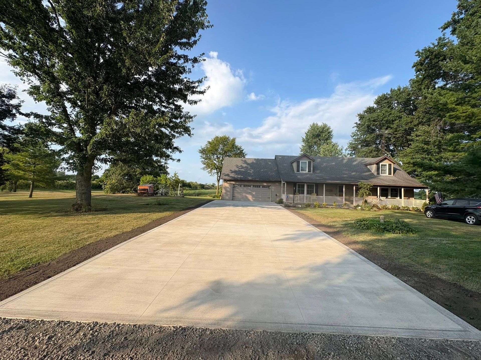 A long, newly poured concrete driveway leads to a single-story ranch-style house surrounded by trees on a sunny day.