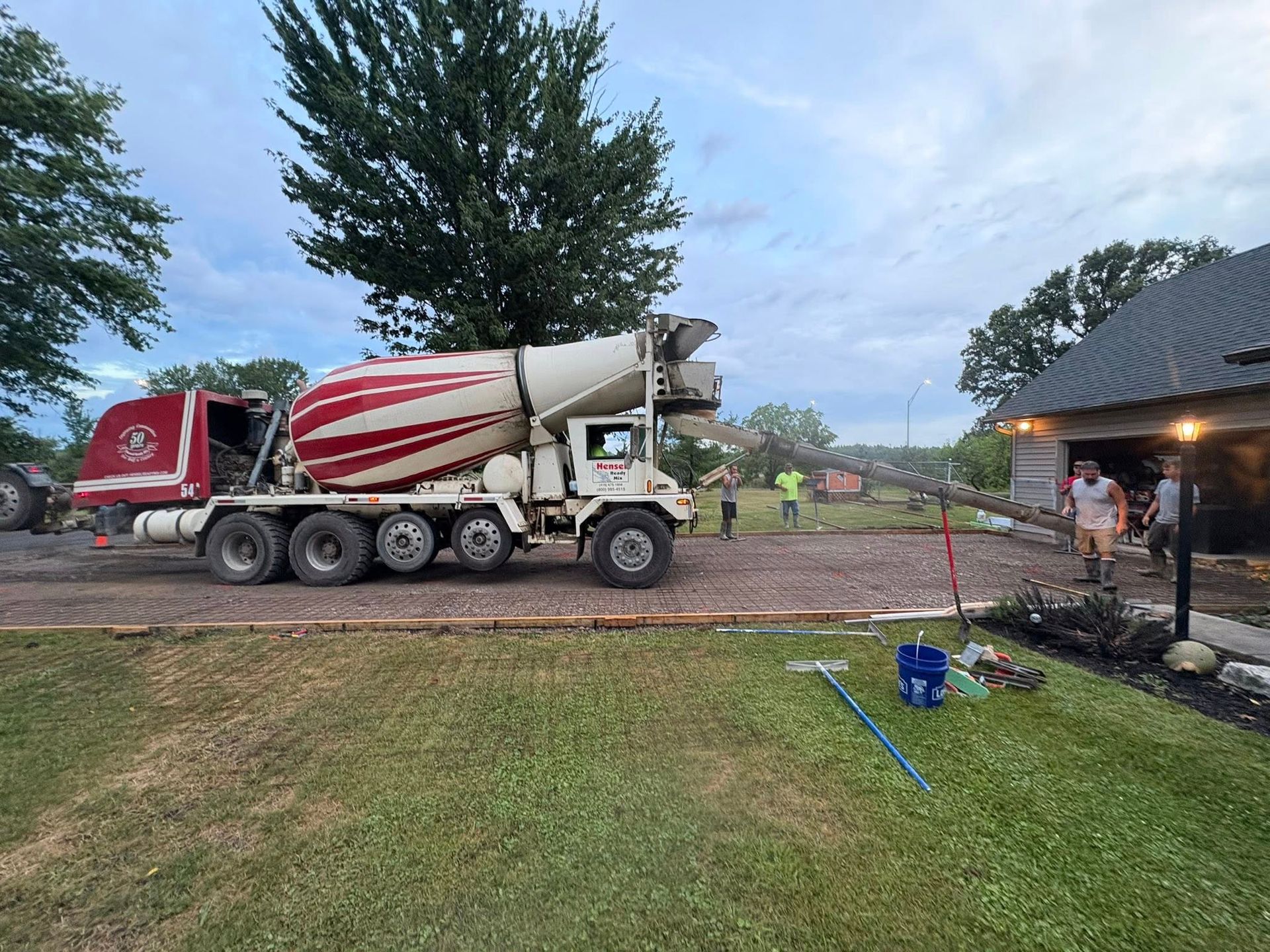 A red and white concrete mixer truck pours concrete into a forms setup for a driveway in front of a residential garage.