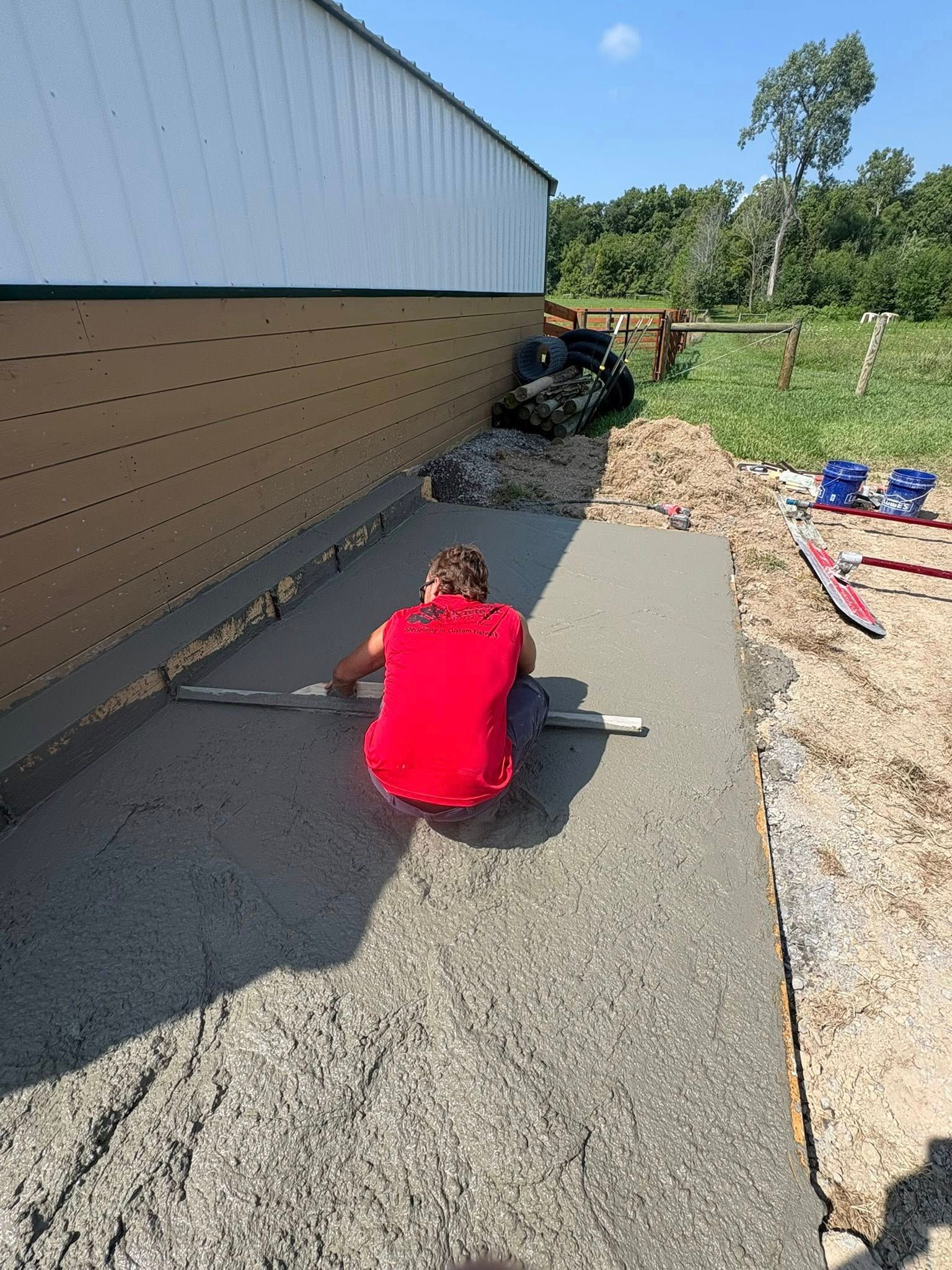 A person in a red shirt kneels on fresh concrete, using a hand tool to smooth the surface beside a building wall.