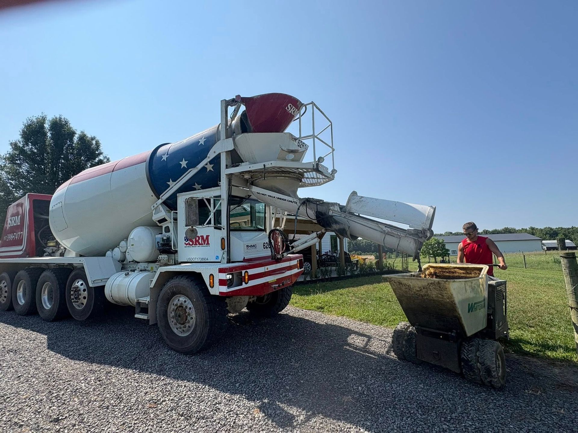 A concrete mixer truck pours wet cement into a small tracked buggy outdoors on a gravel lot under a clear blue sky.