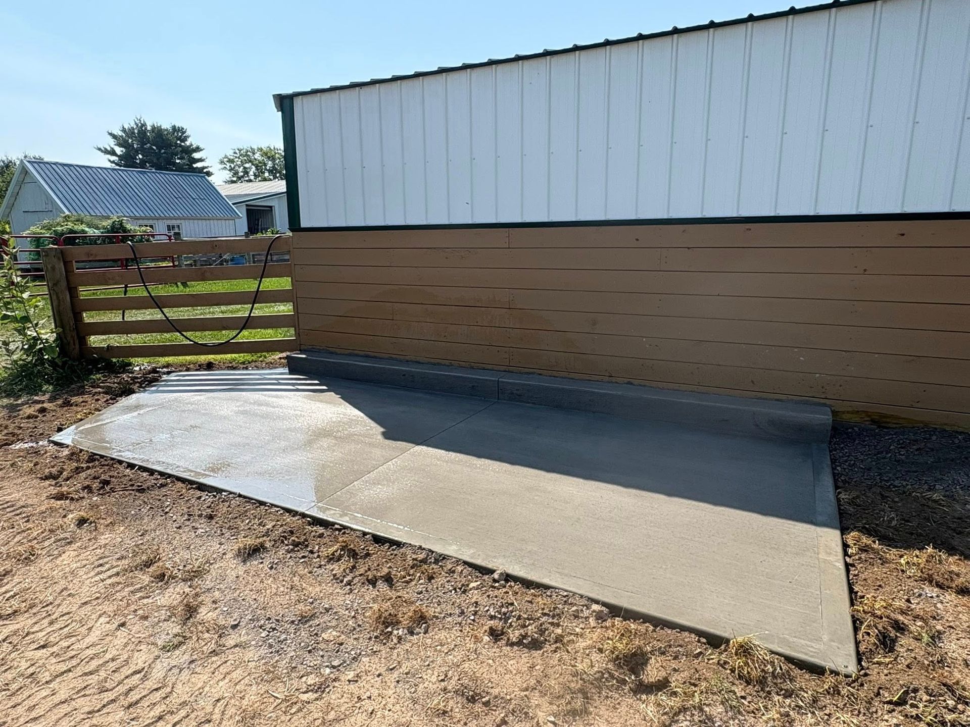 A newly poured concrete pad sits on dirt next to a barn wall, with a wooden fence and another building in the background.