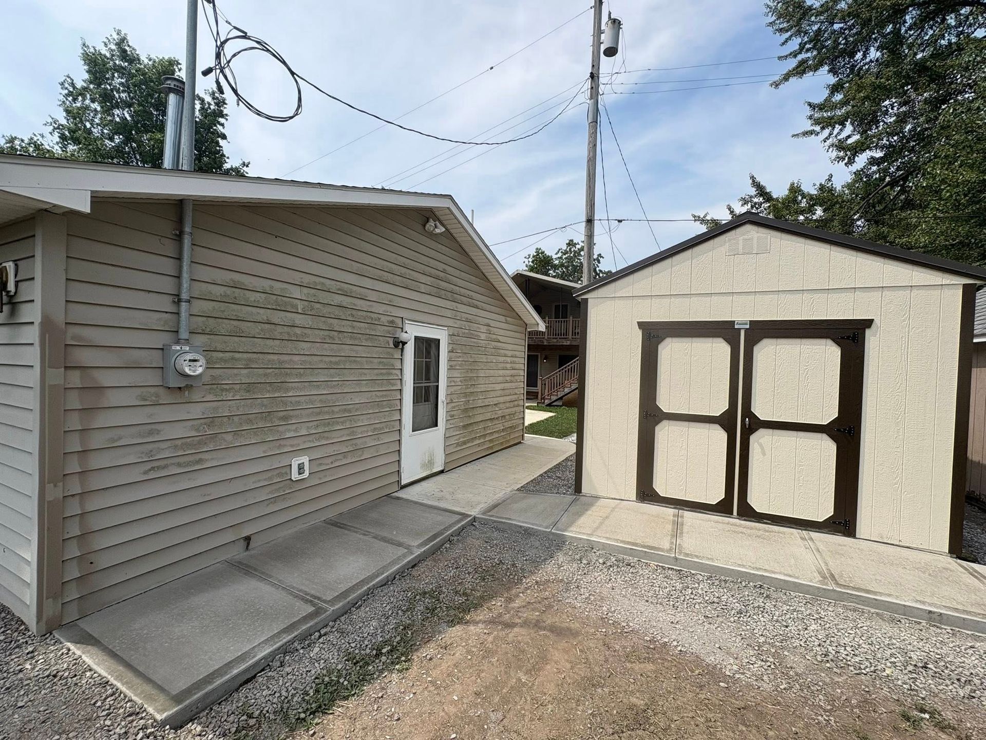 A beige house with a white door and a small, detached shed with brown-trimmed double doors, connected by a concrete path.