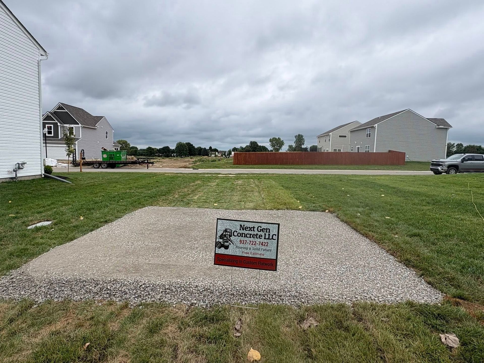 A gravel pad sits in a grassy residential yard with a small sign centered on it, set against a backdrop of new homes.