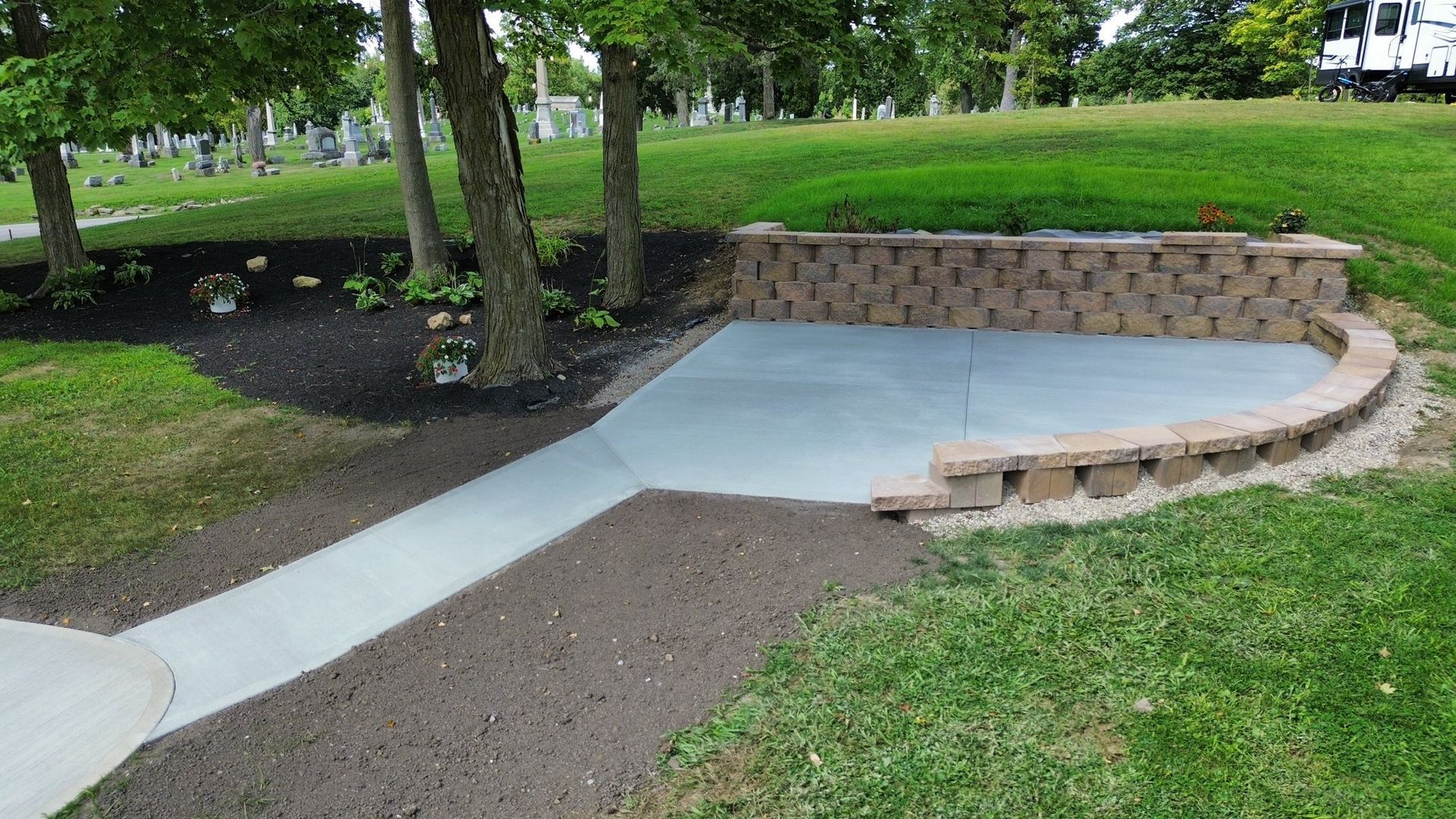 A concrete patio and walkway set in a grassy yard, enclosed by a curved, tiered tan stone retaining wall.