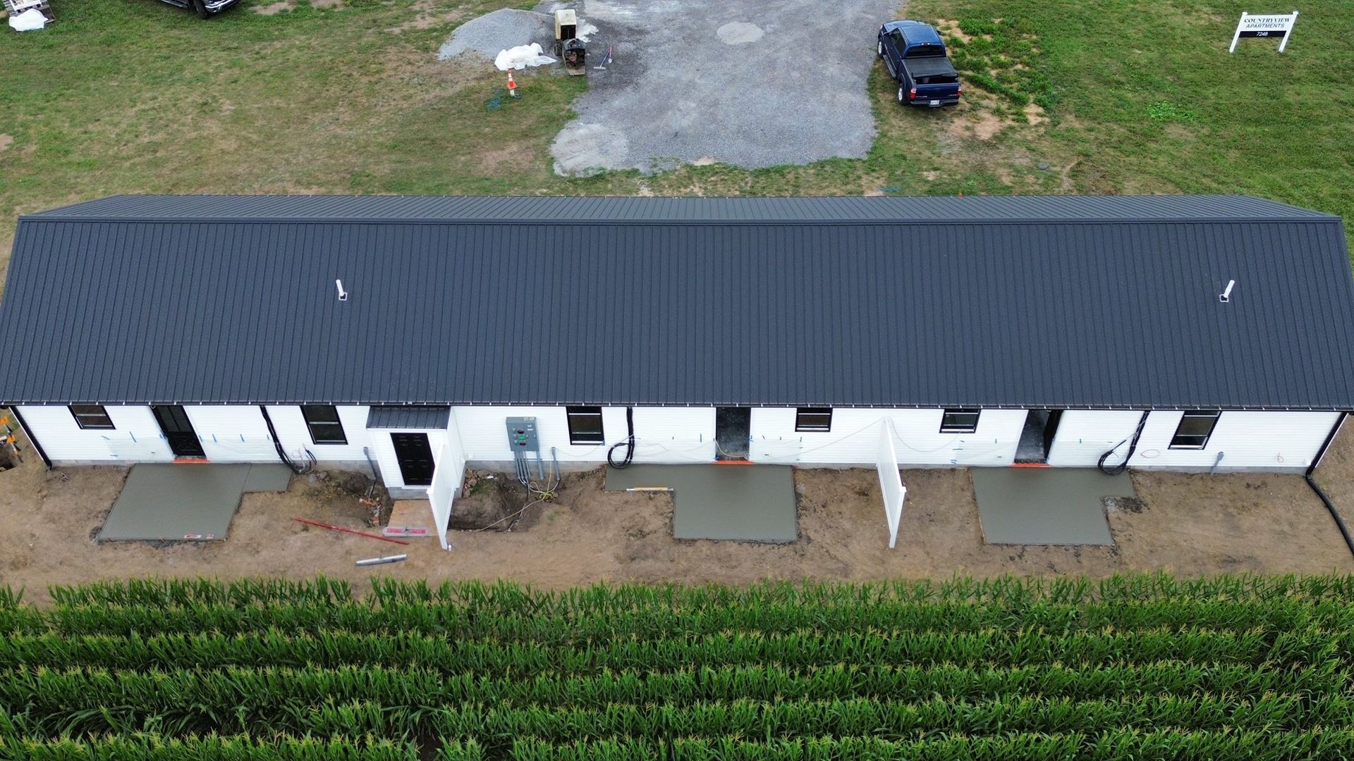 An aerial view of a long, white, multi-unit building with a dark metal roof, located next to a field of crops.