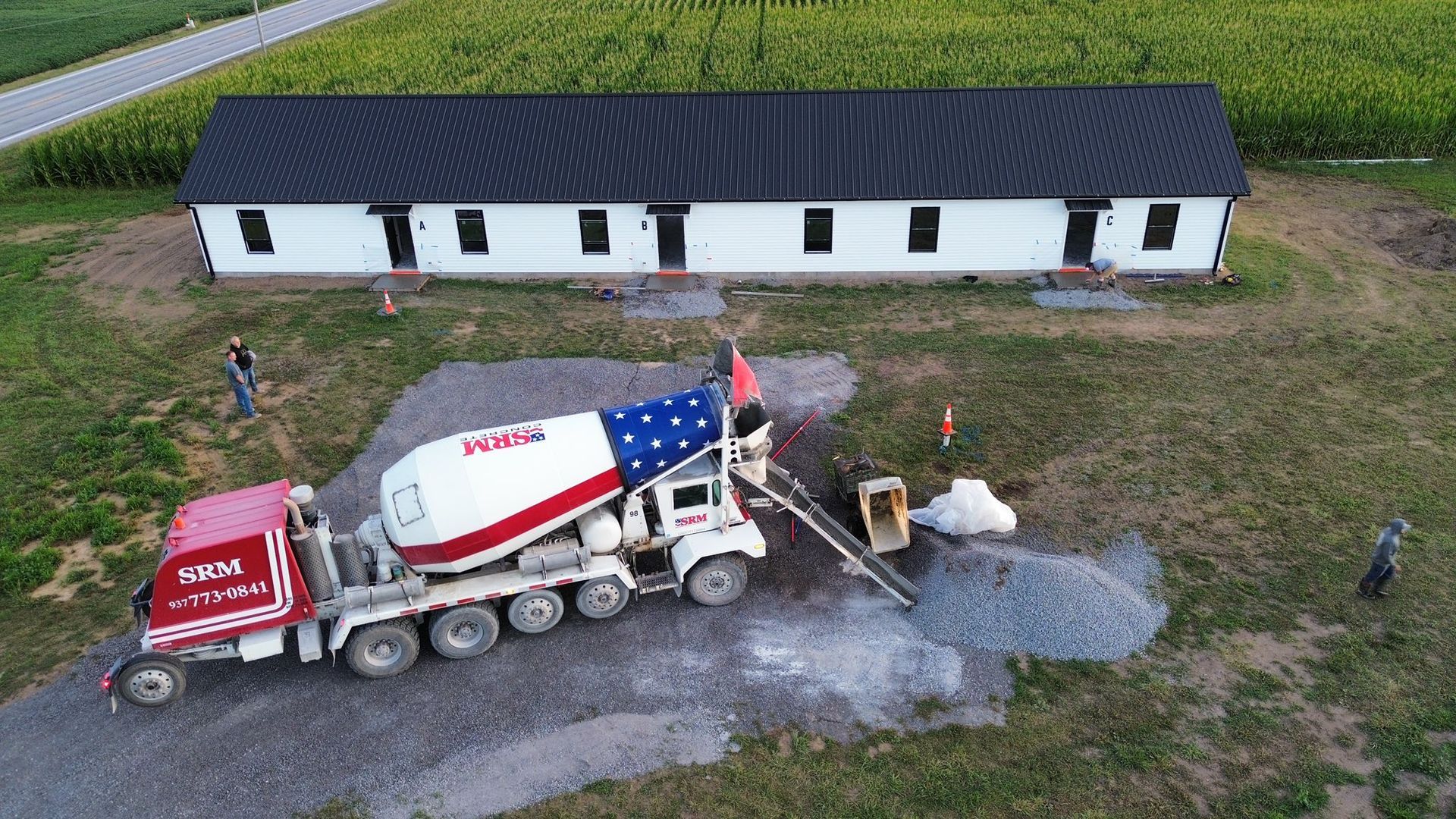 An aerial view shows a red and white cement truck pouring gravel near a long, white building in a rural field.