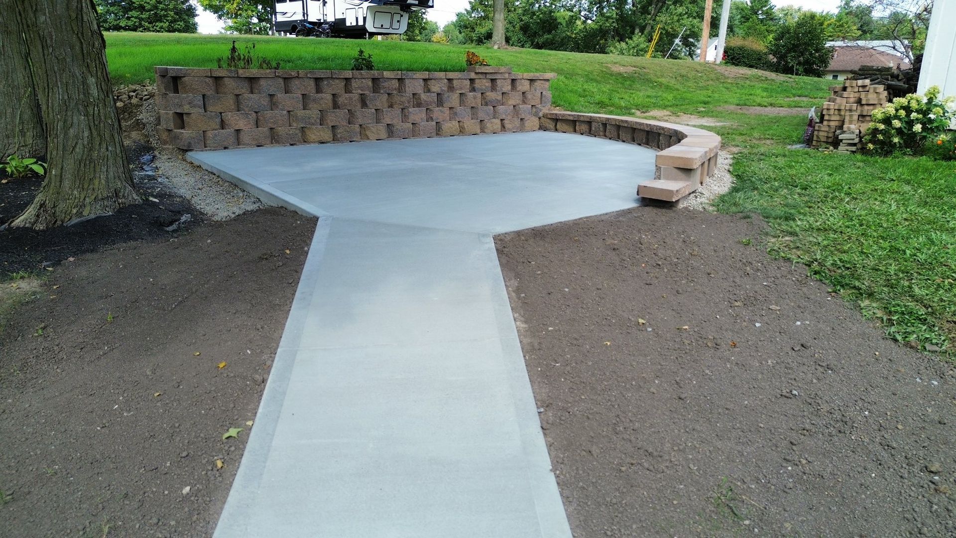 A newly poured gray concrete walkway leading to a semi-circular patio, bordered by a tiered stone retaining wall.