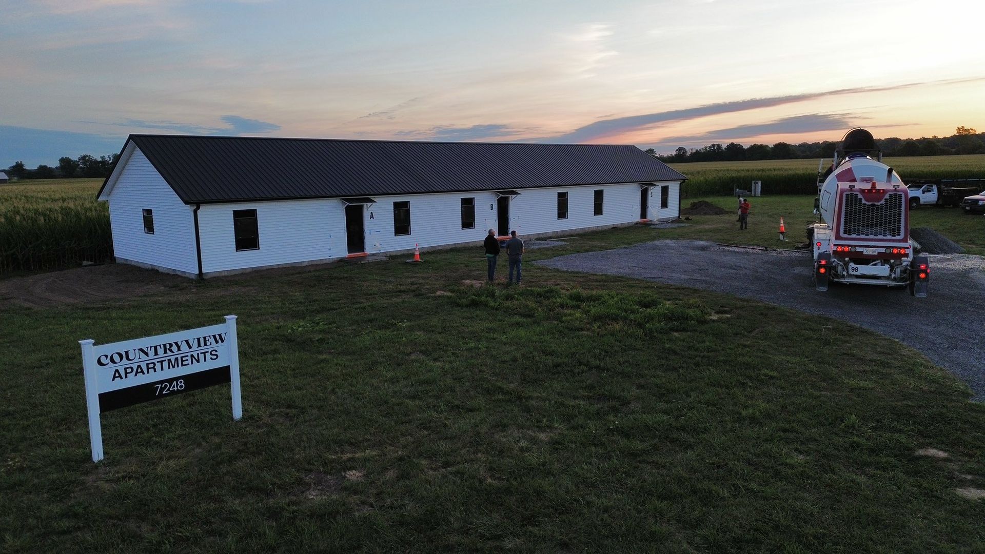 A long, white, single-story building at dusk, with a cement truck parked nearby and a sign in the foreground.