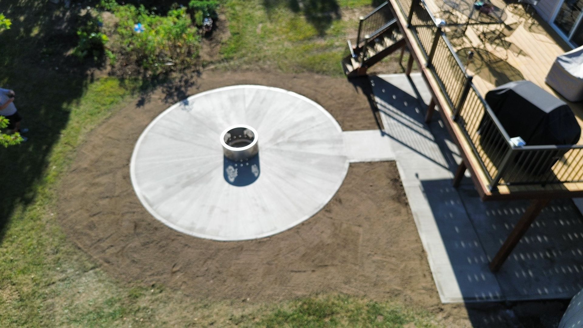 An aerial view of a circular concrete fire pit patio connected by a short walkway to a backyard wooden deck.