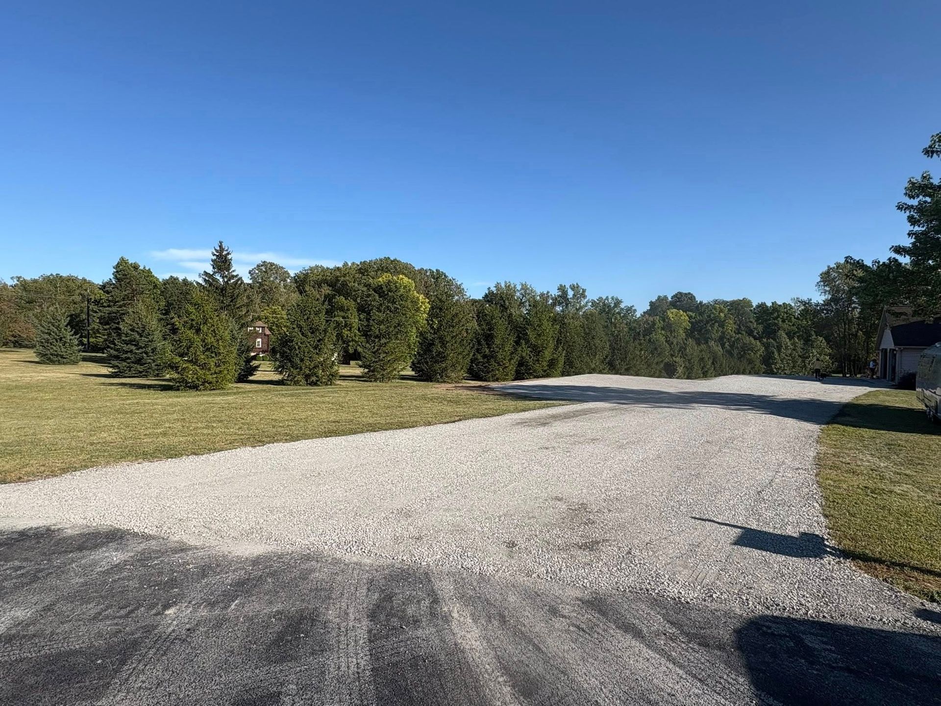 A wide gravel driveway leads toward a line of green trees under a clear, bright blue sky.