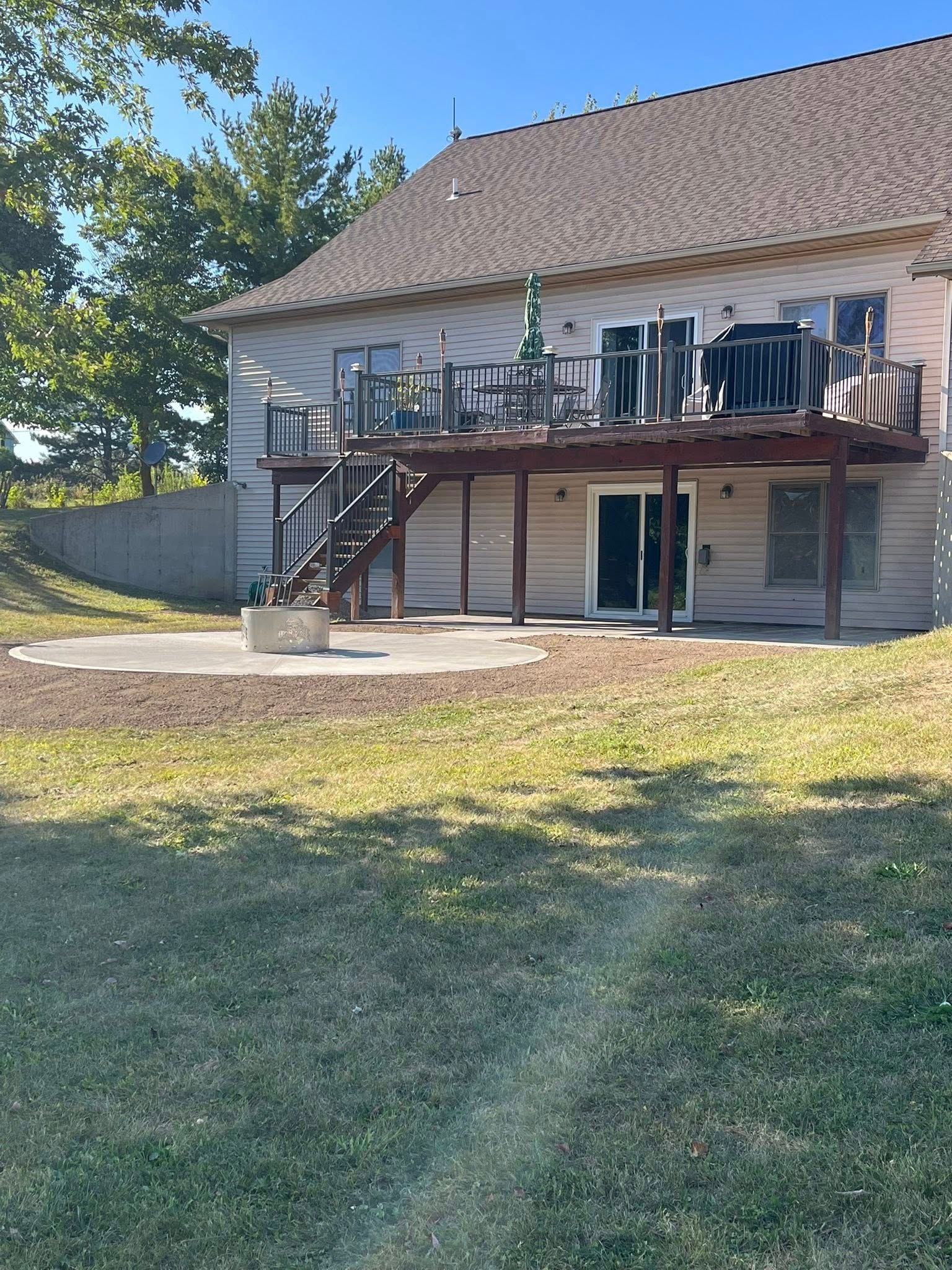 A two-story light-colored house with a dark wooden deck, a staircase, and a concrete patio in the grassy backyard.