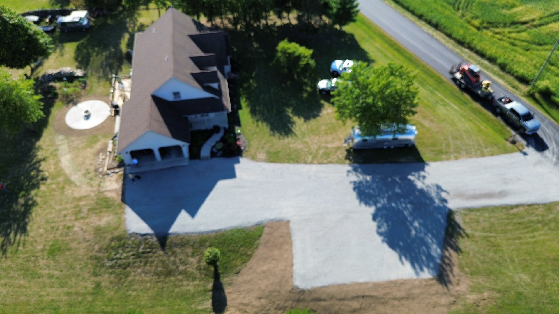 Aerial view of a house with a large paved driveway, surrounded by a grassy lawn, trees, and a road with a parked vehicle.