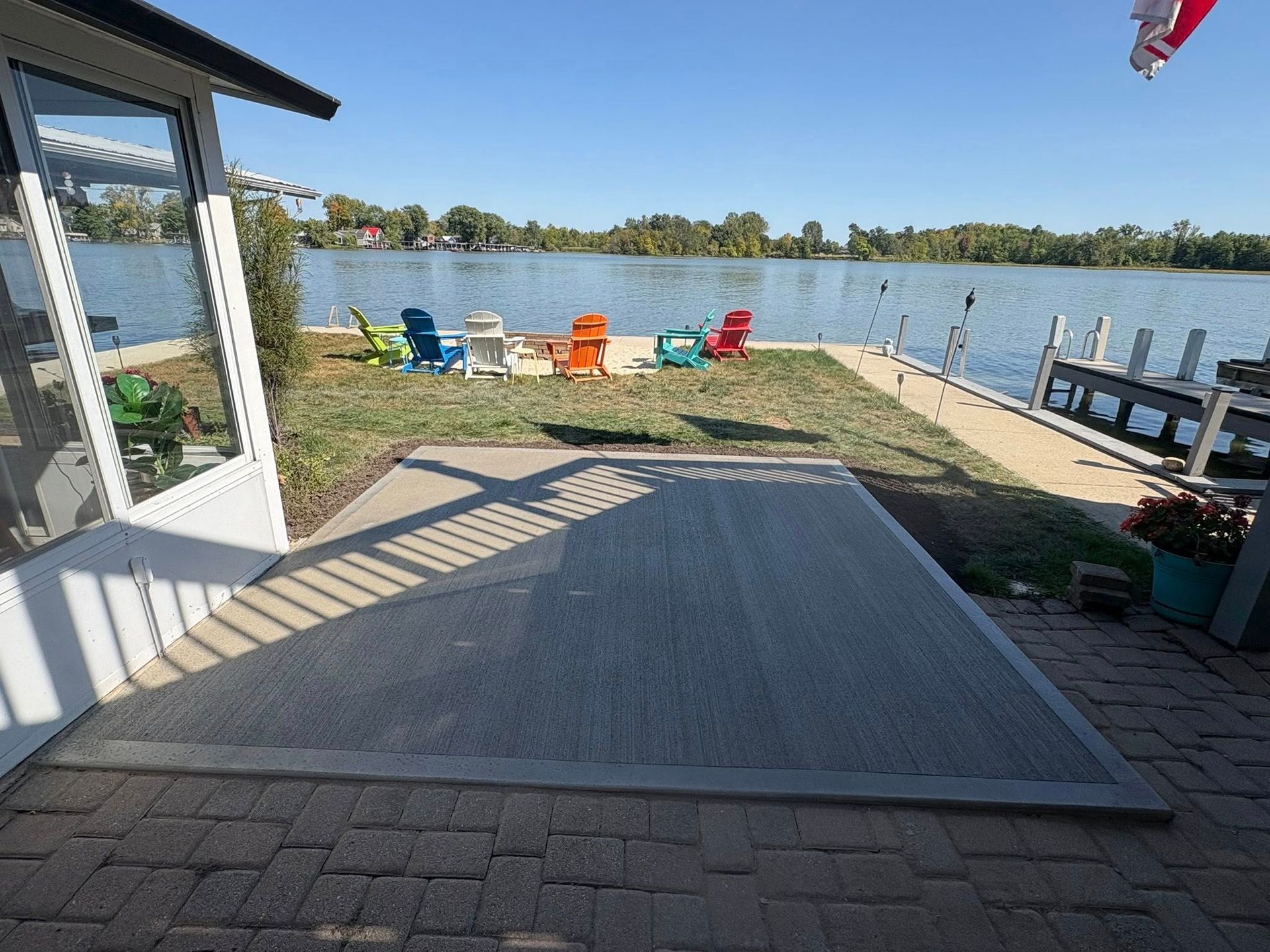 A patio overlooking a lake with several colorful Adirondack chairs on the grass and a wooden dock on the right.