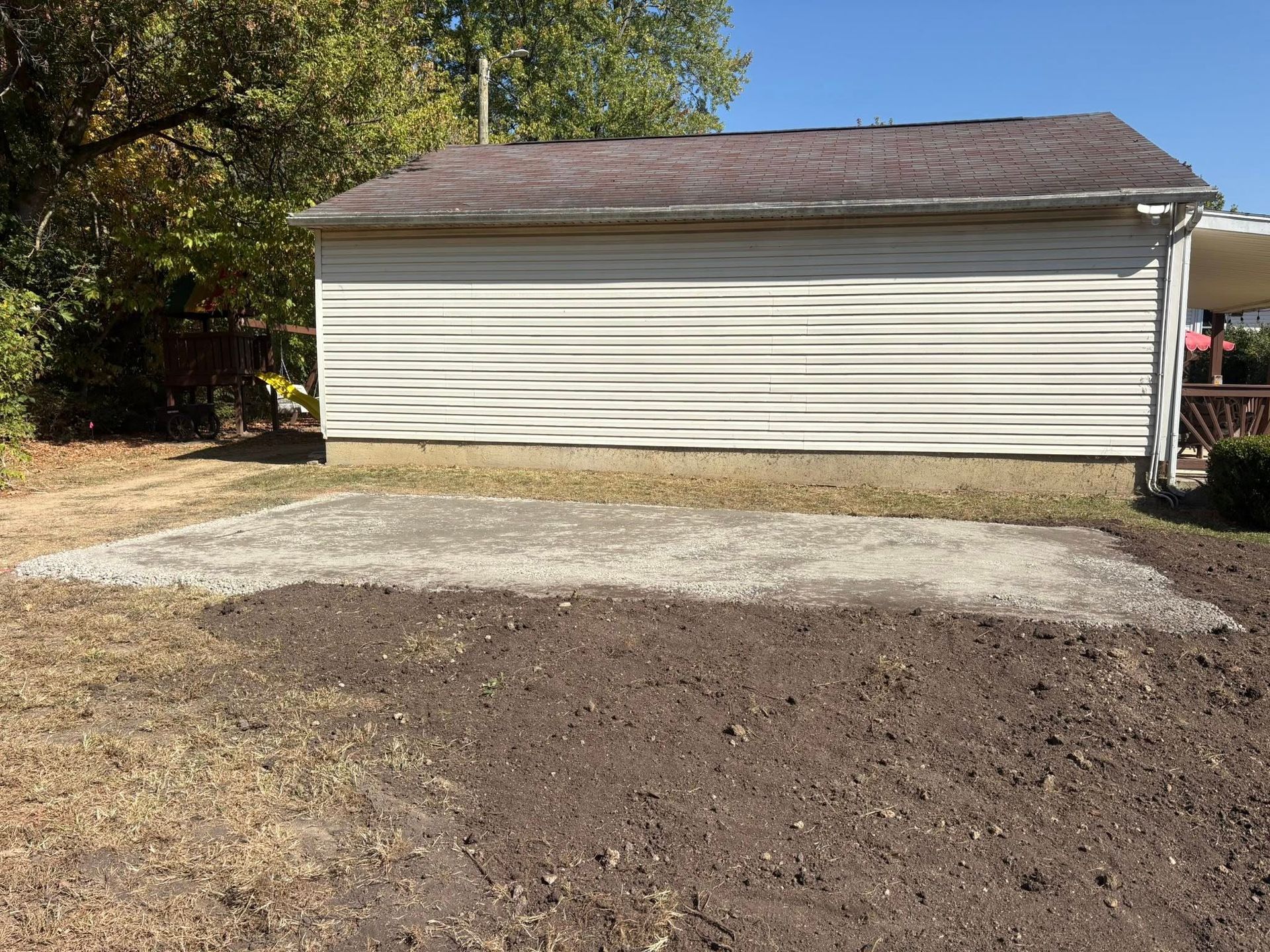A rectangular patch of gravel in a yard sits in front of a white, horizontal-sided garage under a clear blue sky.
