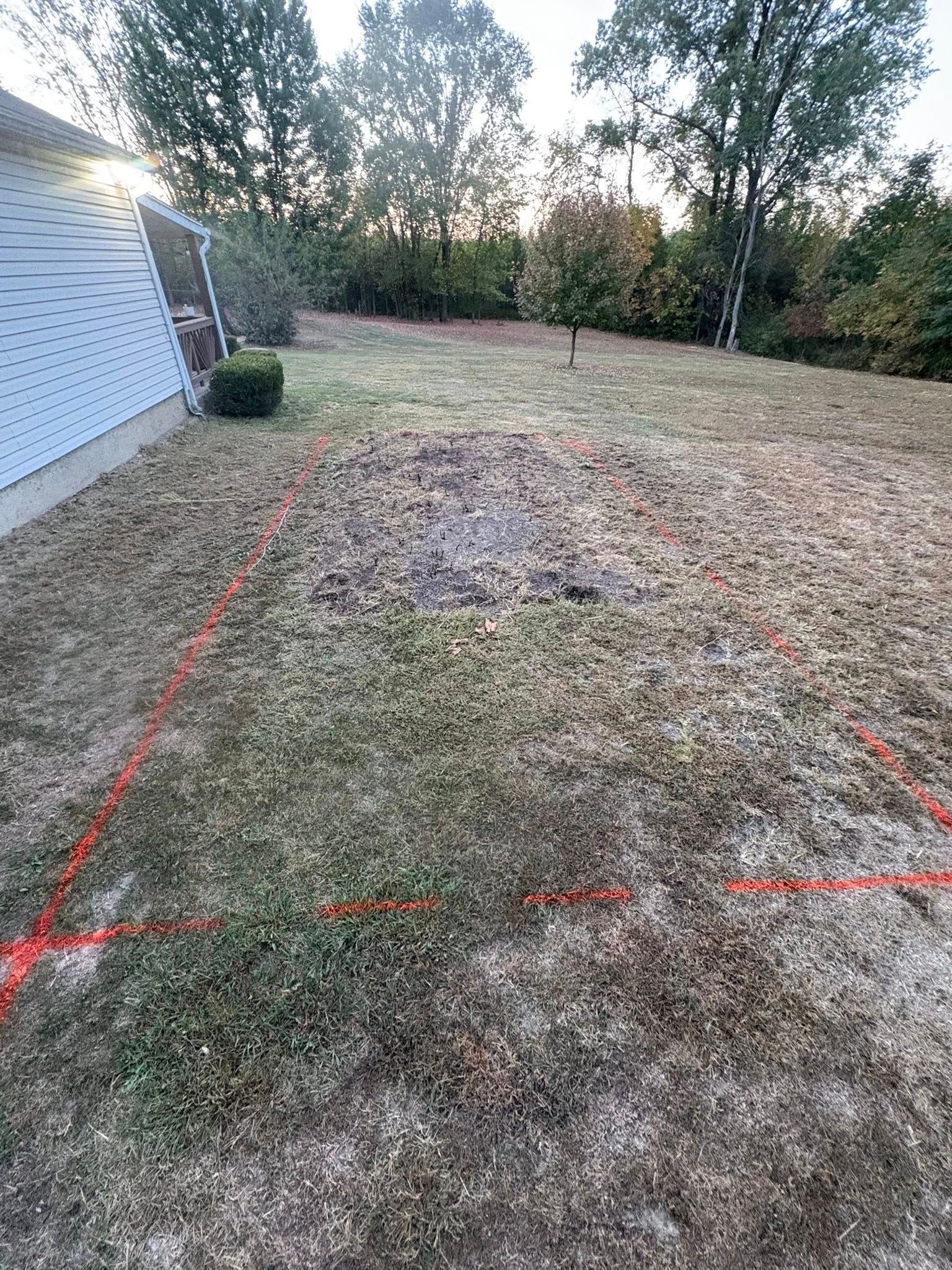 A rectangular area in a grassy backyard marked off with orange string, featuring a patch of dark soil in the center.