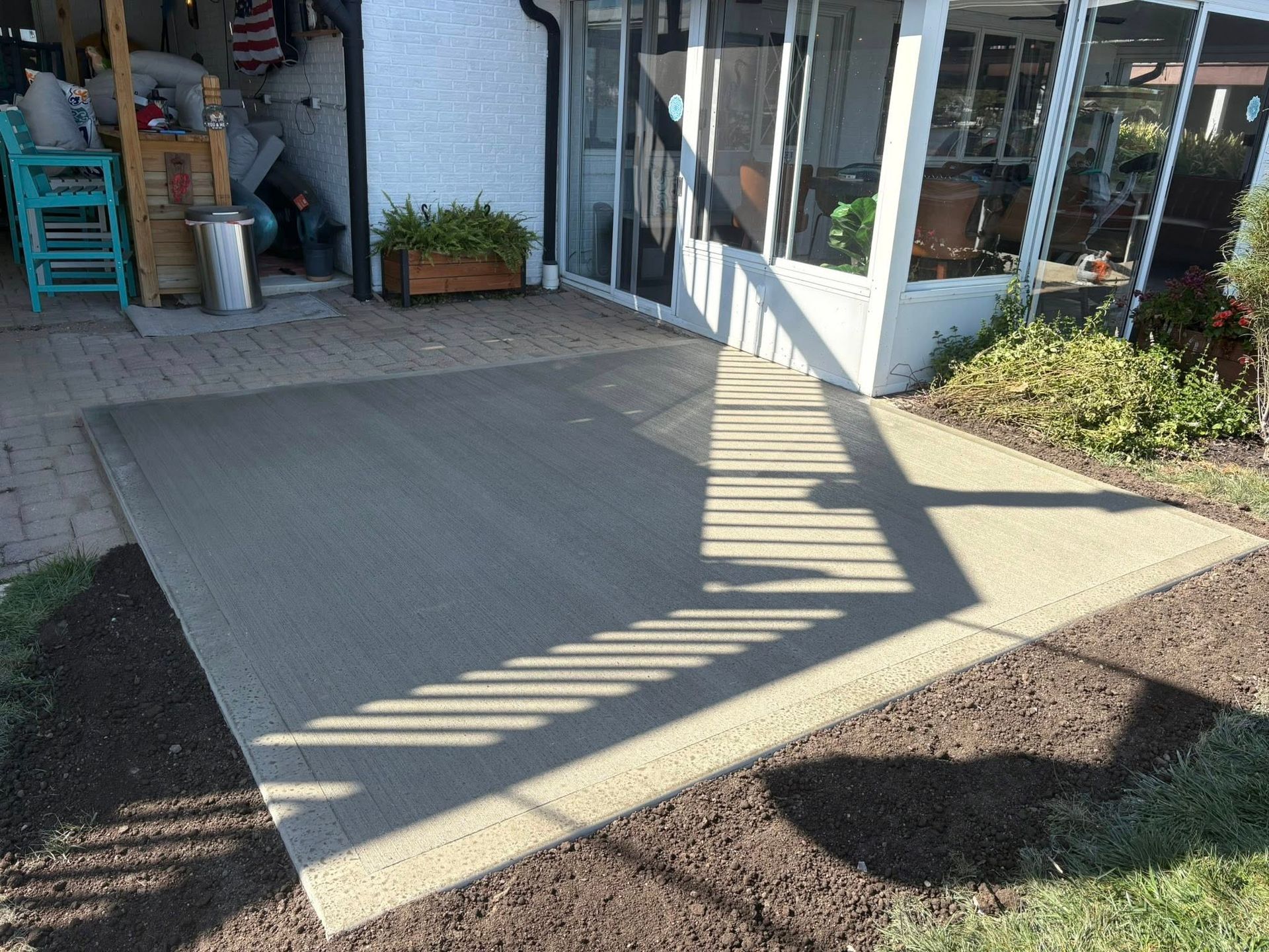 A concrete patio area in a backyard next to a house with a screened-in porch and garden bed.