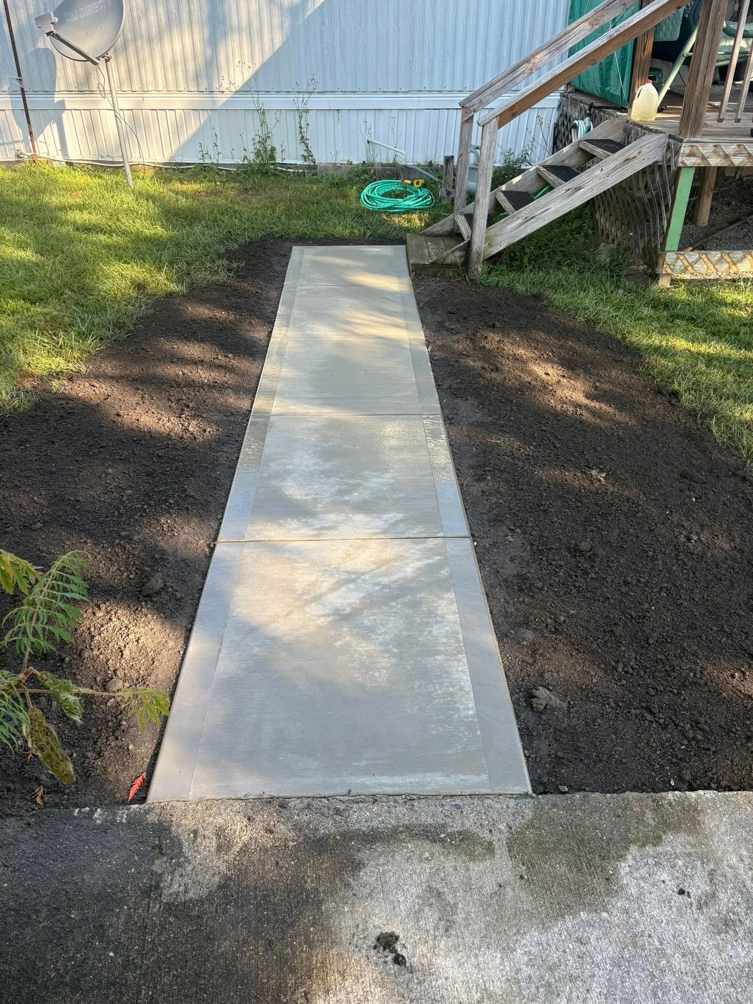 A newly poured concrete walkway leads from a gravel area through dark mulch toward wooden stairs leading to a building.