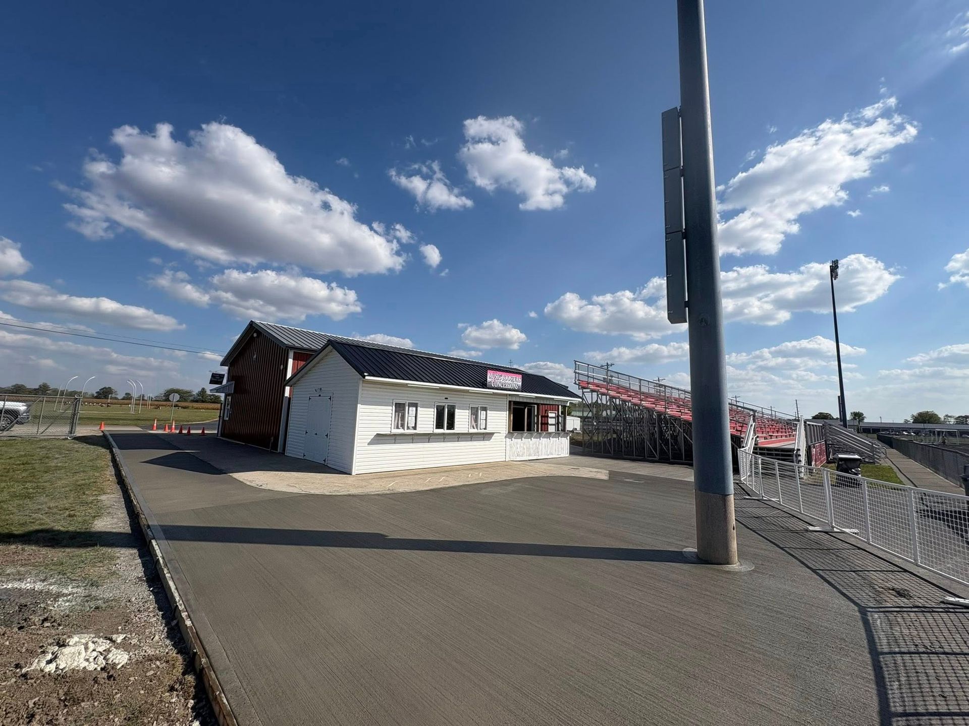 A white concession stand with a striped entrance sits near metal stadium bleachers under a sunny, partly cloudy sky.
