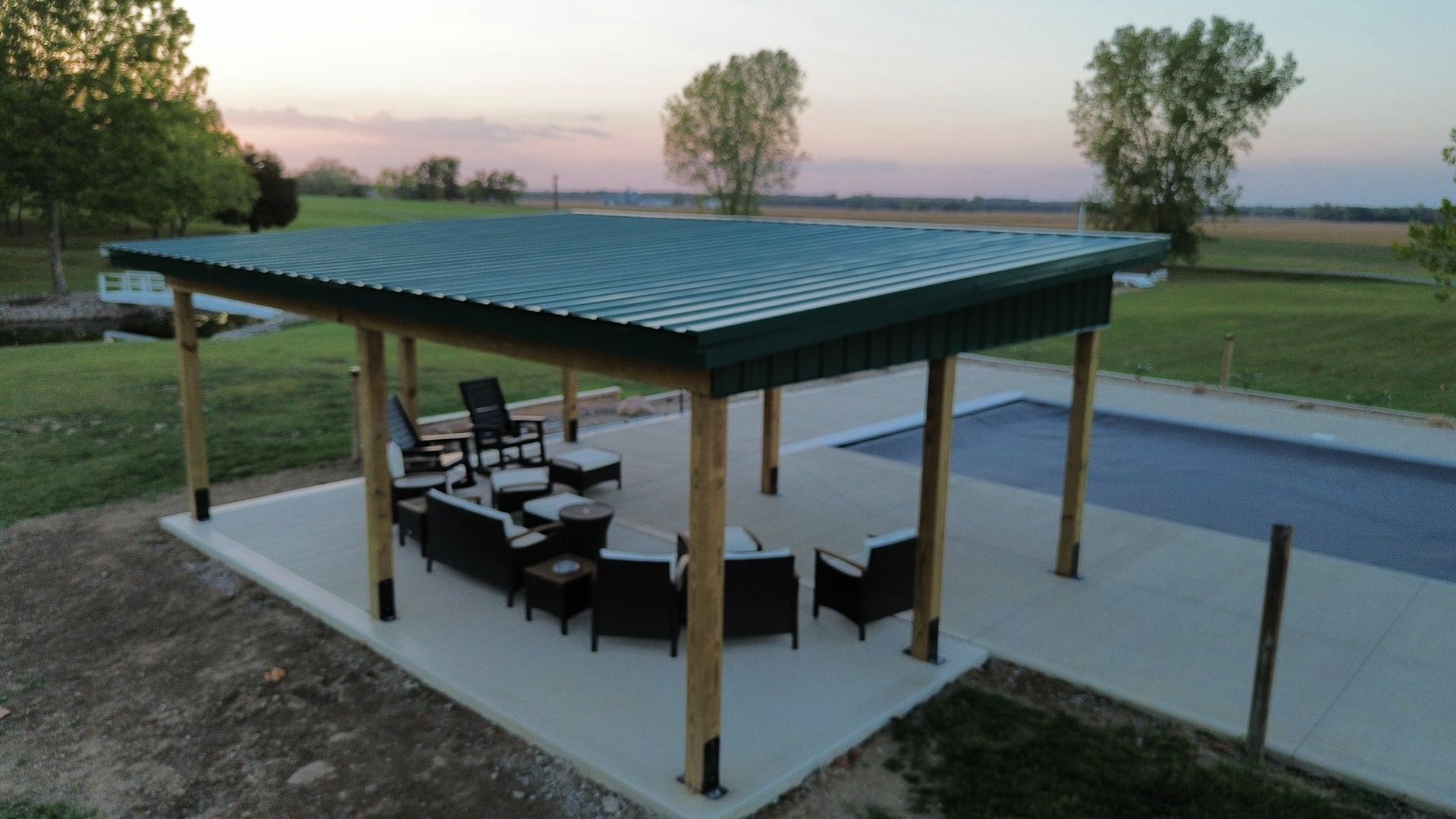 A green-roofed wooden pavilion with patio furniture sits on a concrete pad next to a backyard swimming pool at sunset.
