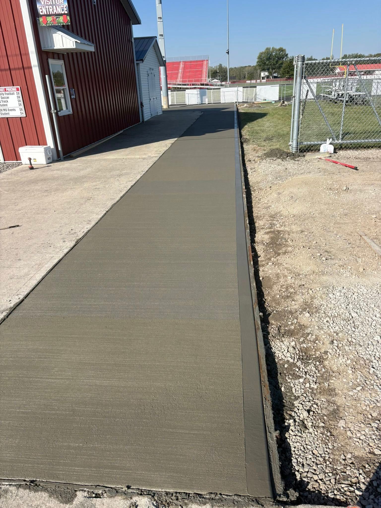 A freshly poured concrete sidewalk stretches away from a red building, bordered by gravel on both sides.