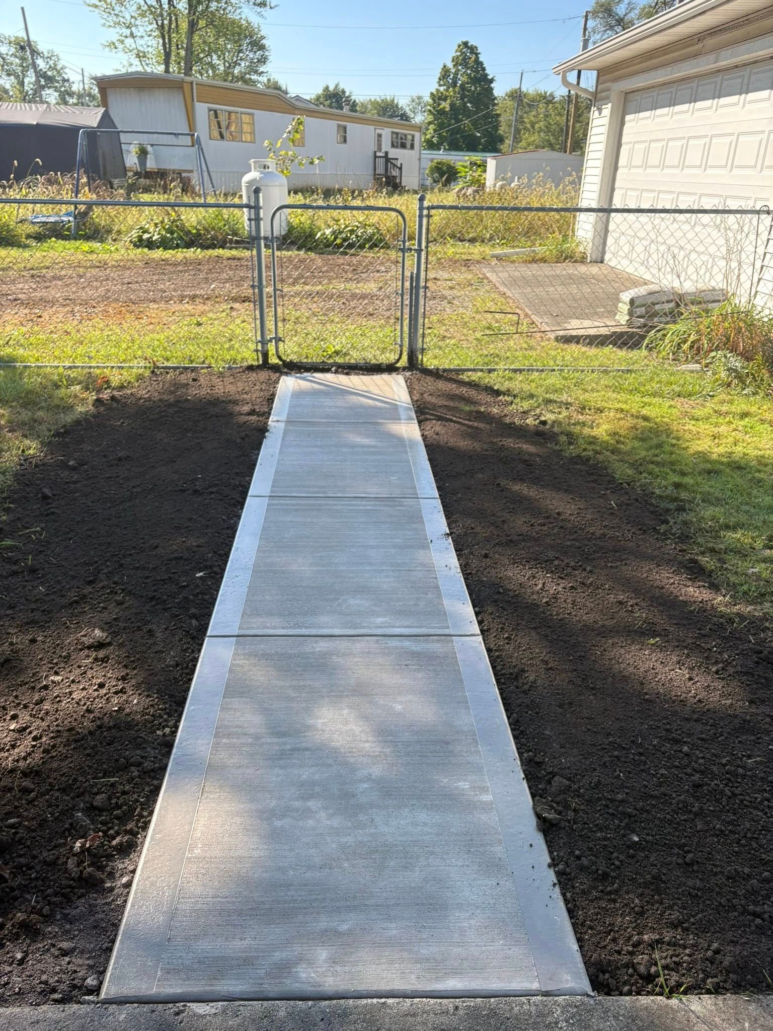 A straight concrete walkway leading toward a metal gate and chain-link fence, flanked by patches of dark, fresh soil.