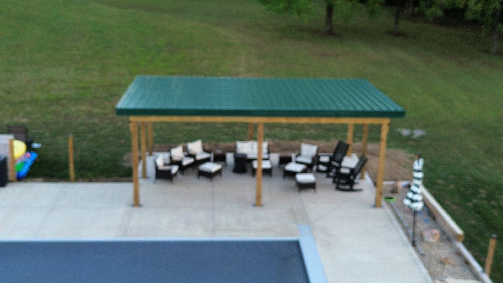 An outdoor patio area with a green-roofed wooden pavilion, furnished with patio chairs and ottomans, next to a pool.