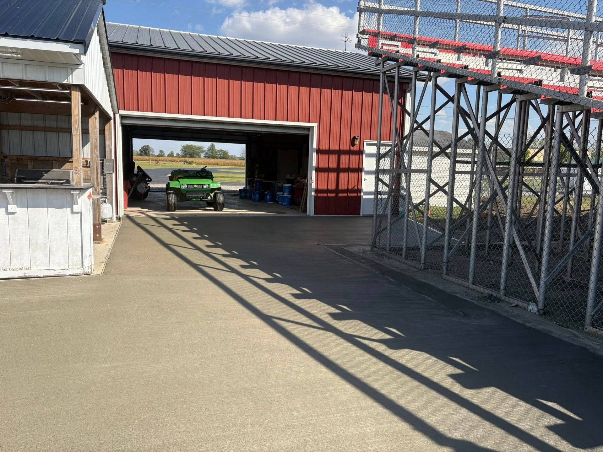 A green utility vehicle sits inside an open-air red barn, viewed from a paved area next to metal stadium bleachers.