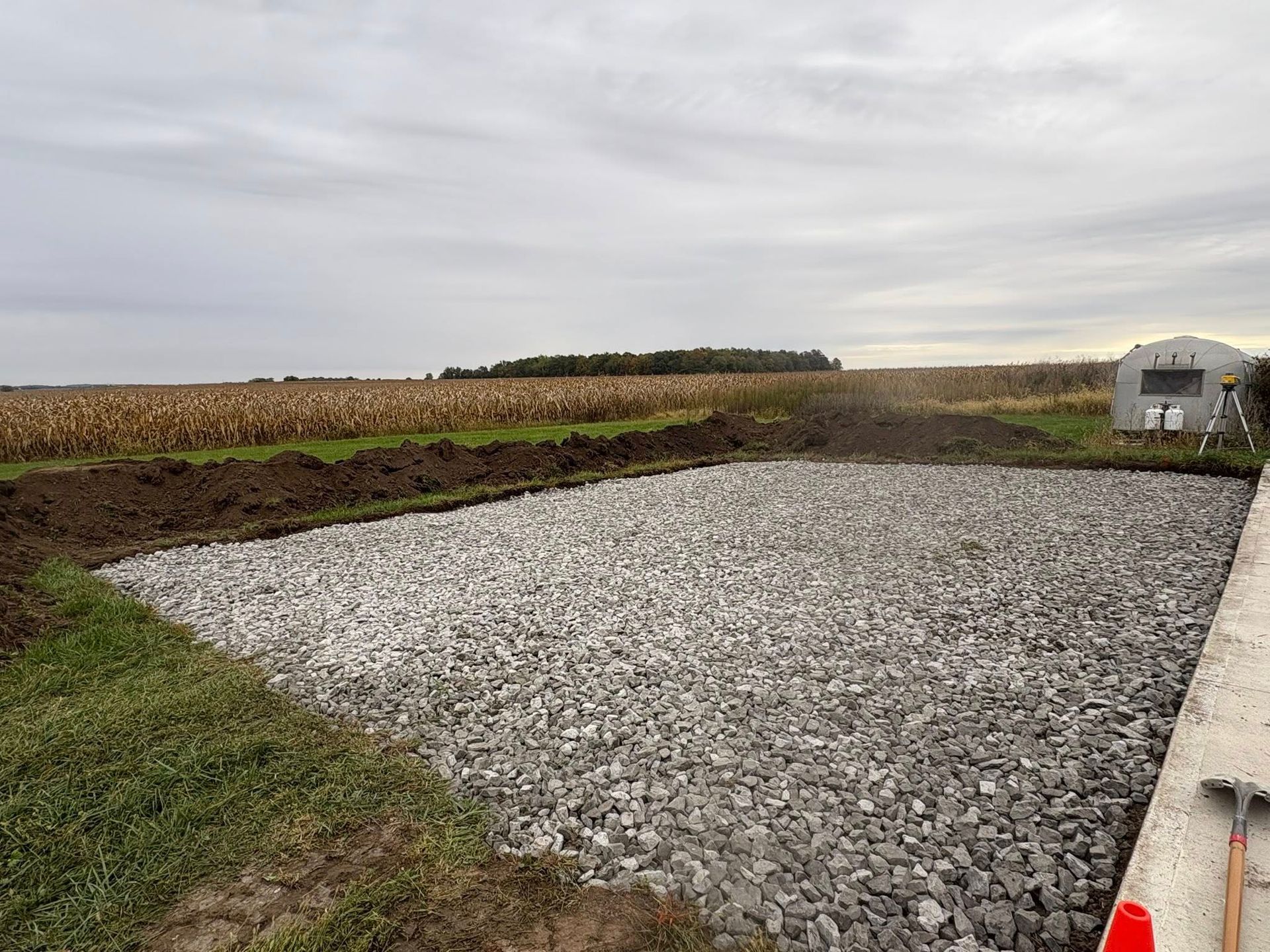 A rectangular gravel patch sits in a grassy field next to a concrete edge, with a cornfield and cloudy sky in the background.