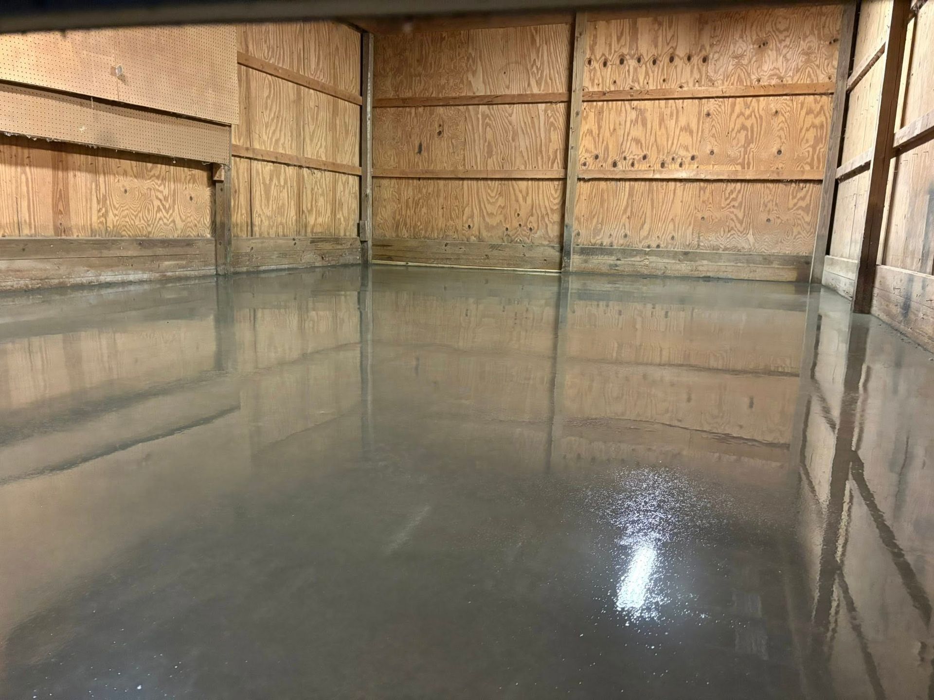 A freshly poured, smooth, gray concrete floor inside a structure with wooden plywood walls.