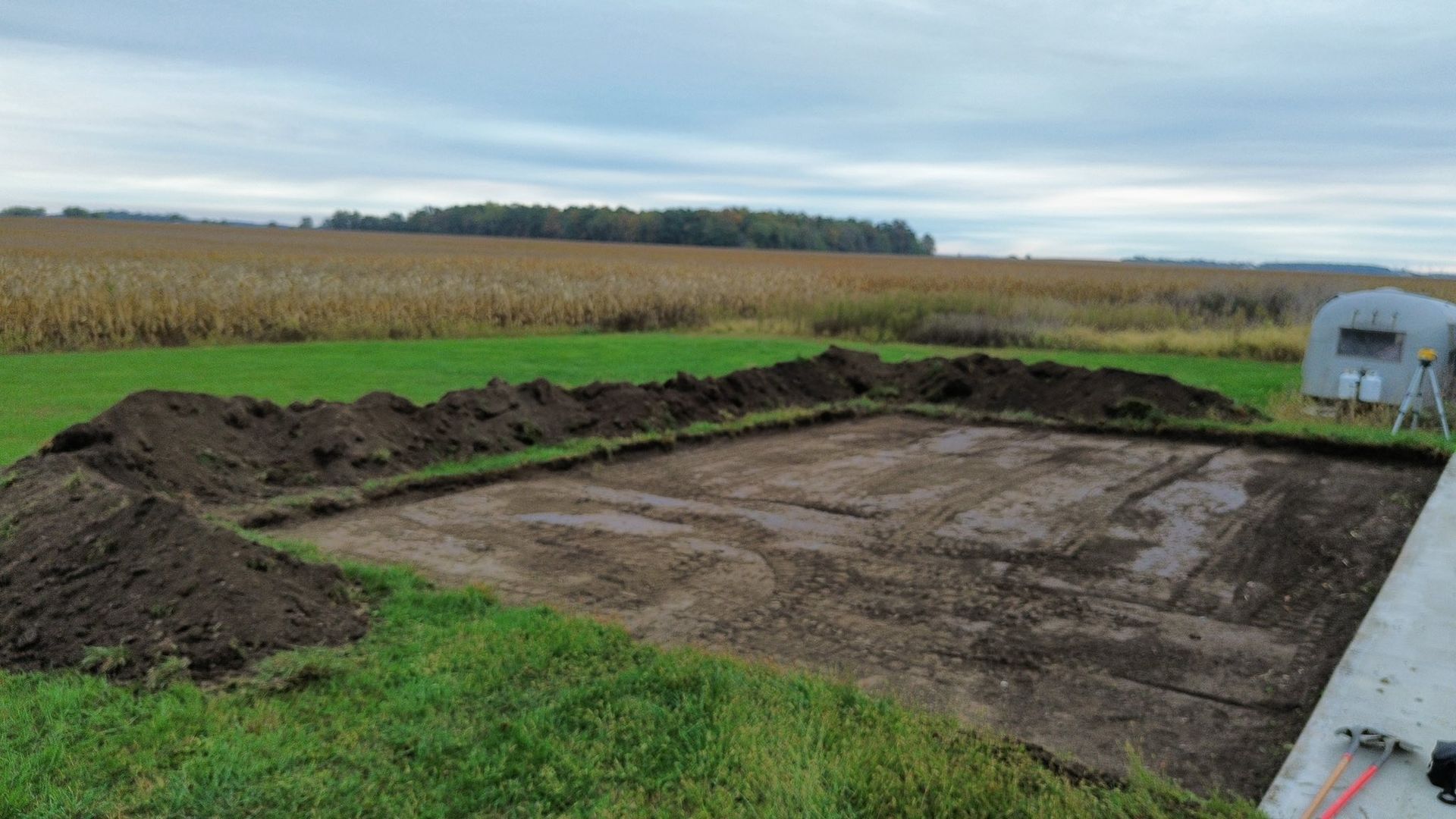 A rectangular patch of excavated soil bordered by piles of dirt, situated in a green field before a harvested cornfield.