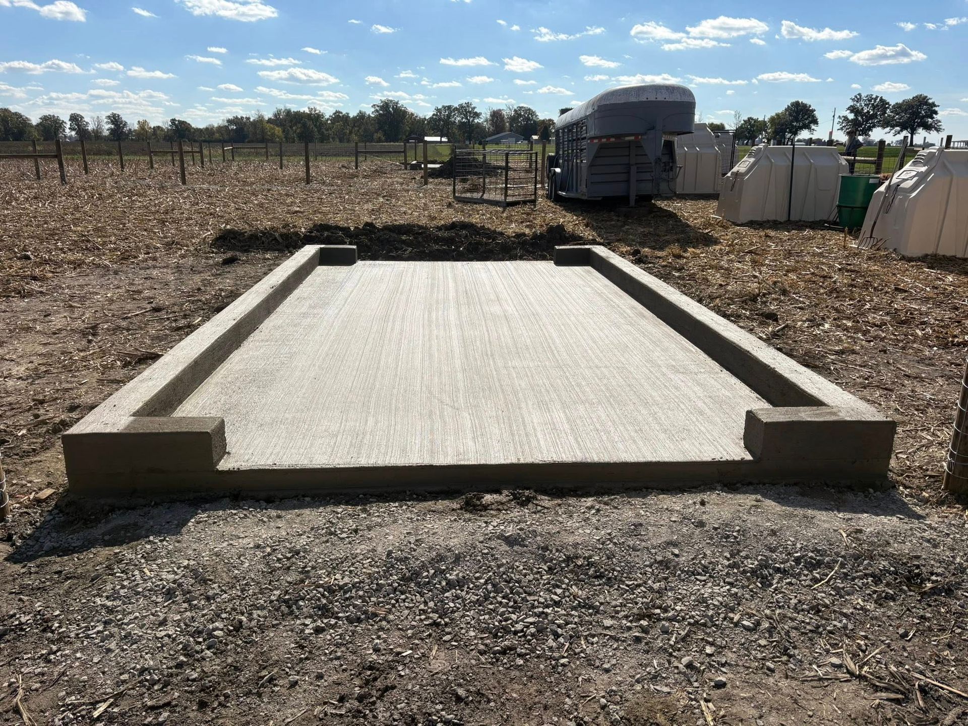 A newly poured concrete slab foundation with raised edges sits in a dirt field under a bright, sunny sky.