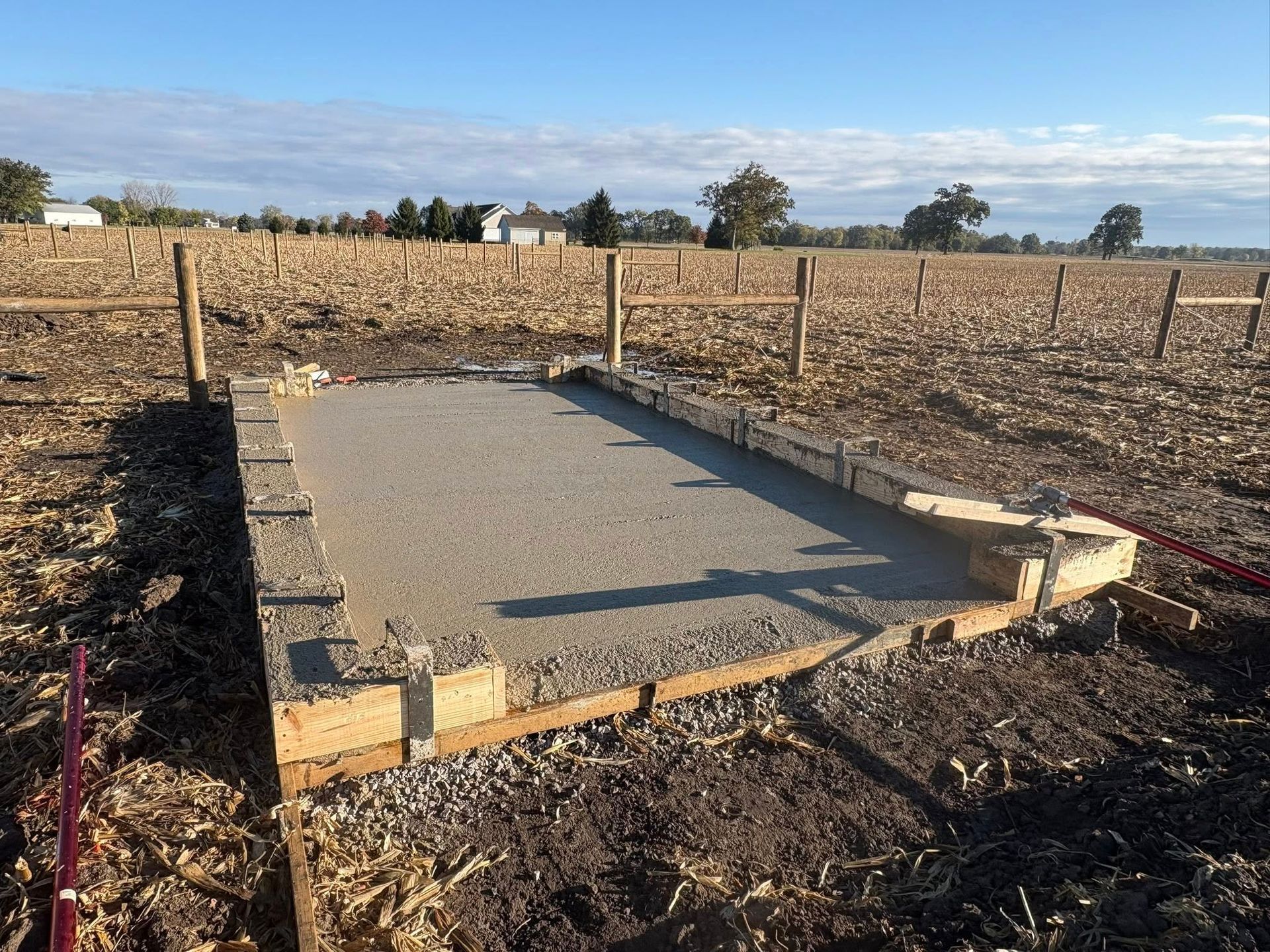 A wooden rectangular form filled with gravel sits in a harvested field under a clear blue sky.