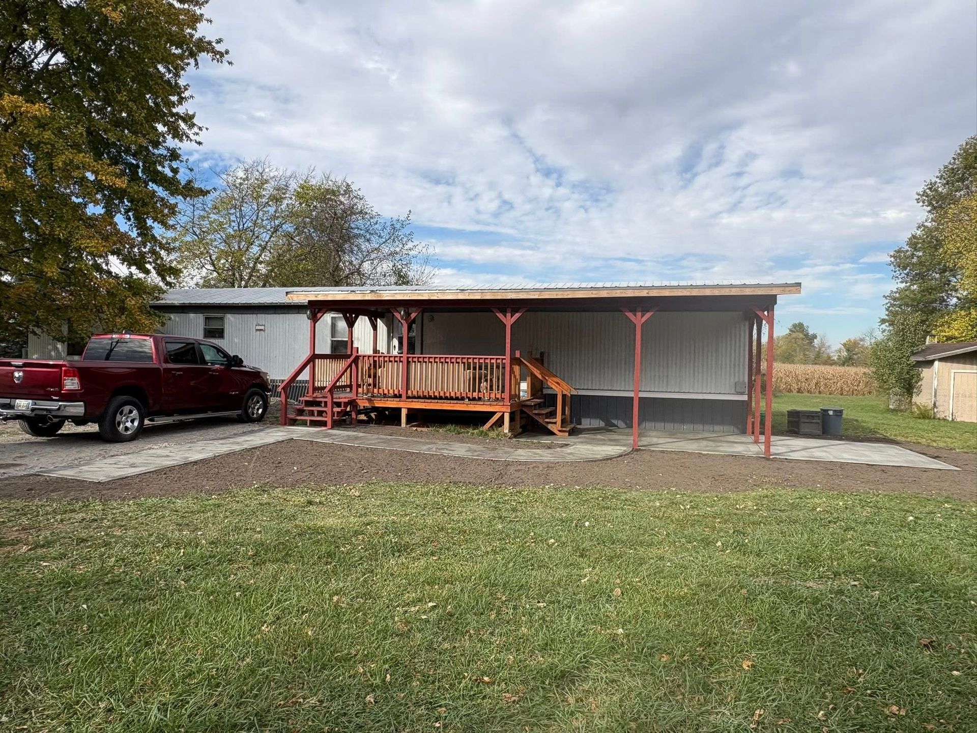 A red pickup truck is parked in front of a white mobile home with a covered wooden porch under a cloudy sky.