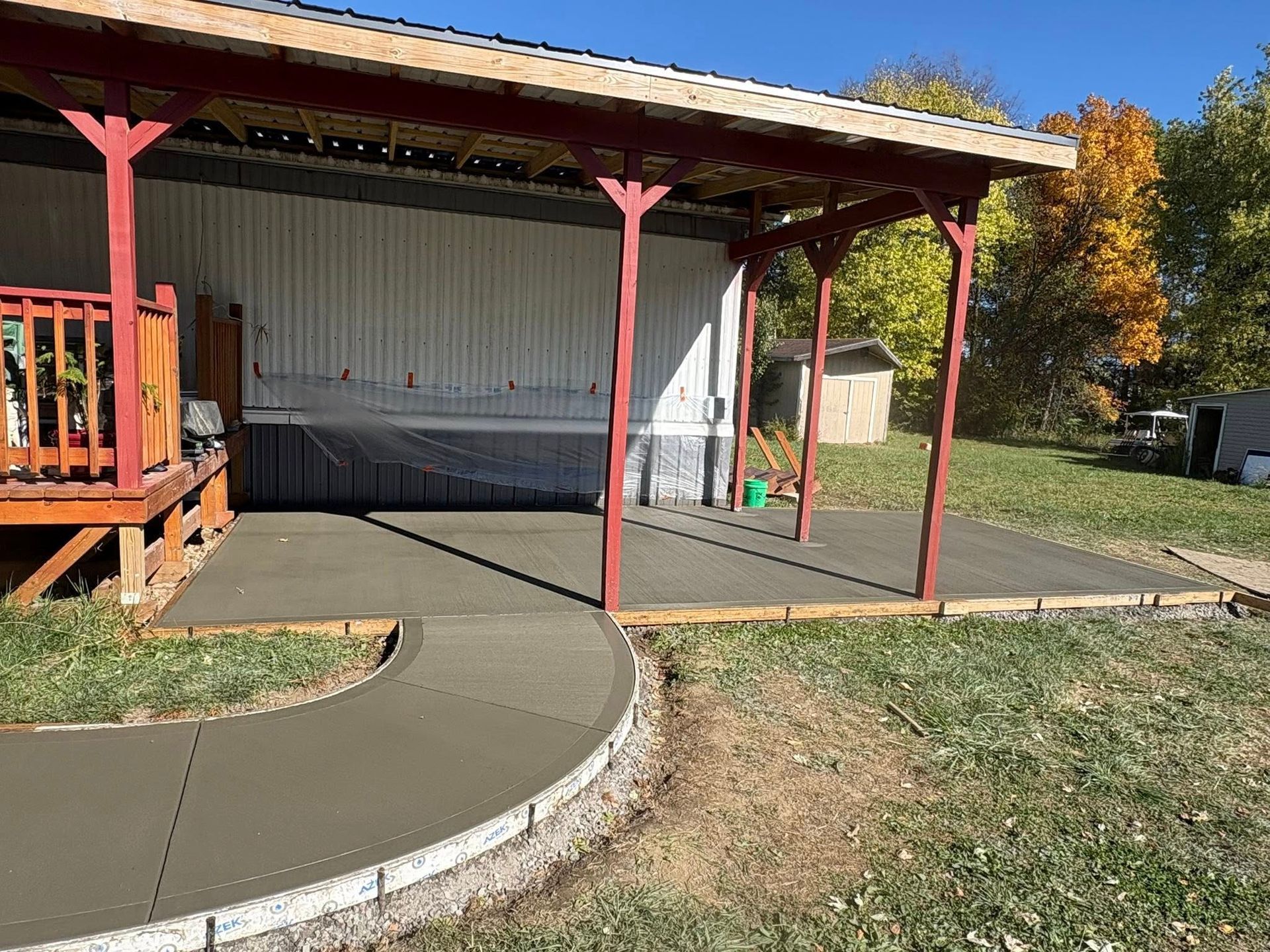Freshly poured concrete patio and walkway under a red-framed wooden roof structure next to a building.
