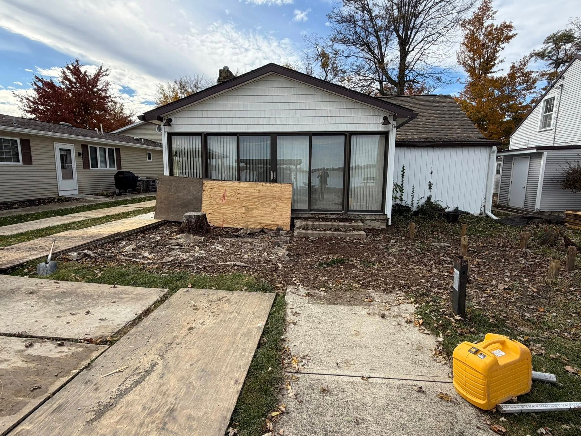 A small, single-story house with white siding, partially boarded windows, a concrete driveway, and a yellow portable pump.