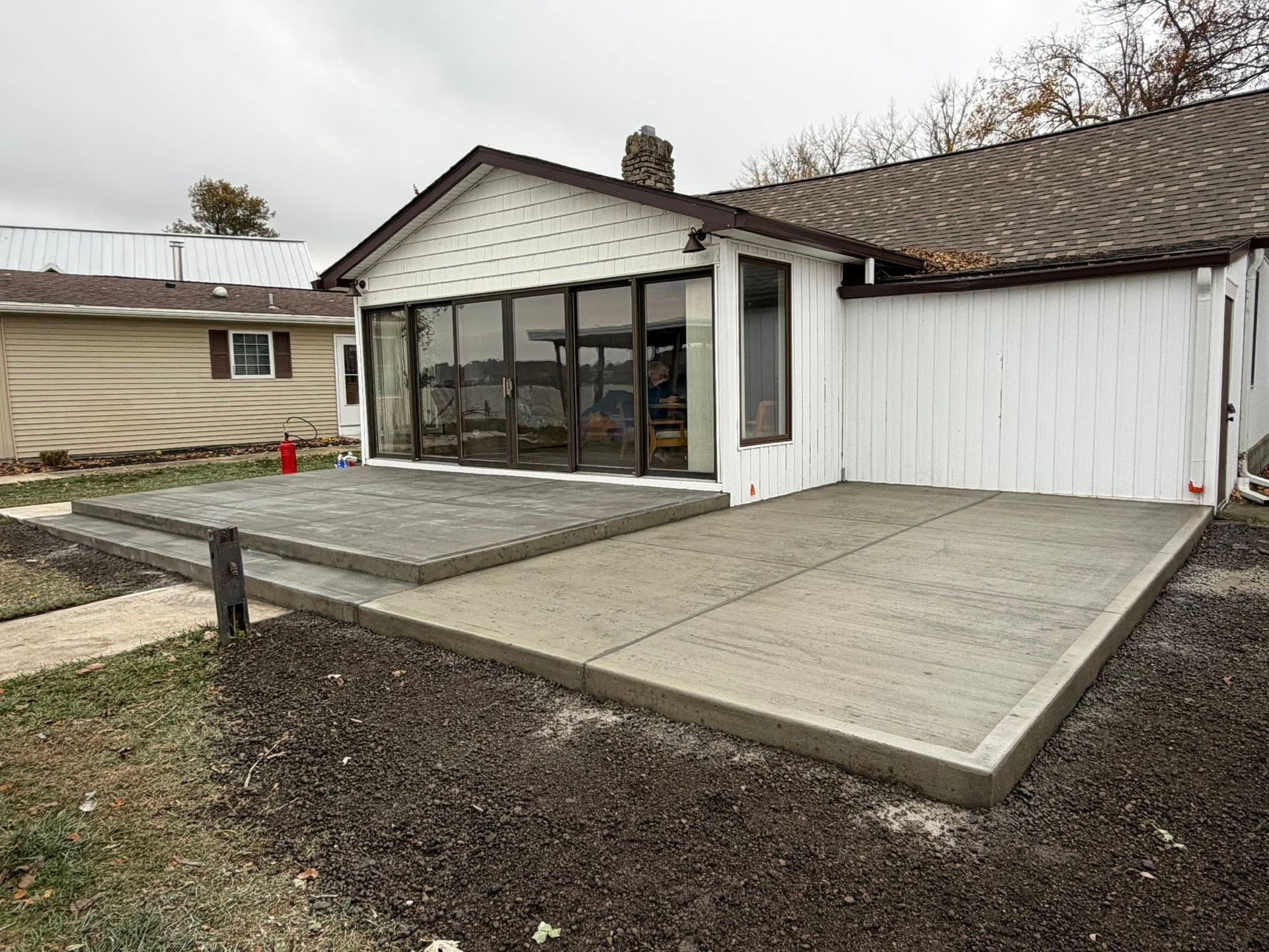 A newly poured concrete patio attached to a house with white siding and glass sliding doors, set against a lakeside yard.