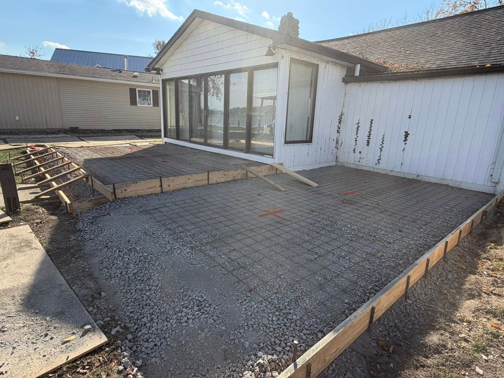 A house exterior under renovation with a wooden-framed gravel base for a patio, prepared for pouring concrete.