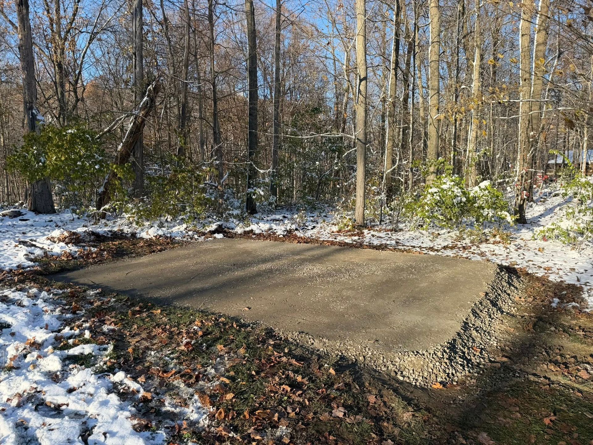 A rectangular gravel shed foundation surrounded by snow and trees in a wooded area.