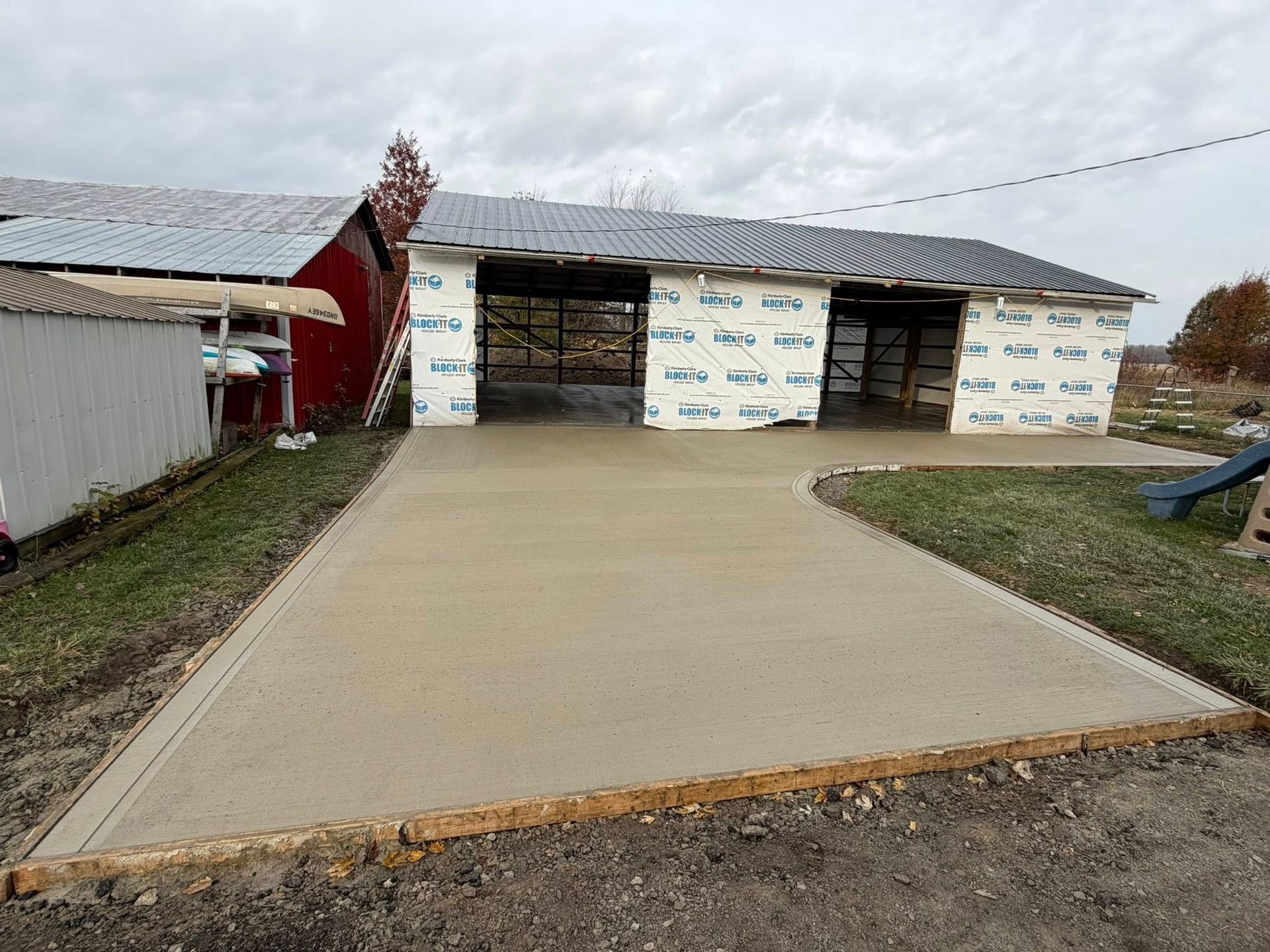 A freshly poured concrete driveway leads to a pole barn with white insulation panels under construction.