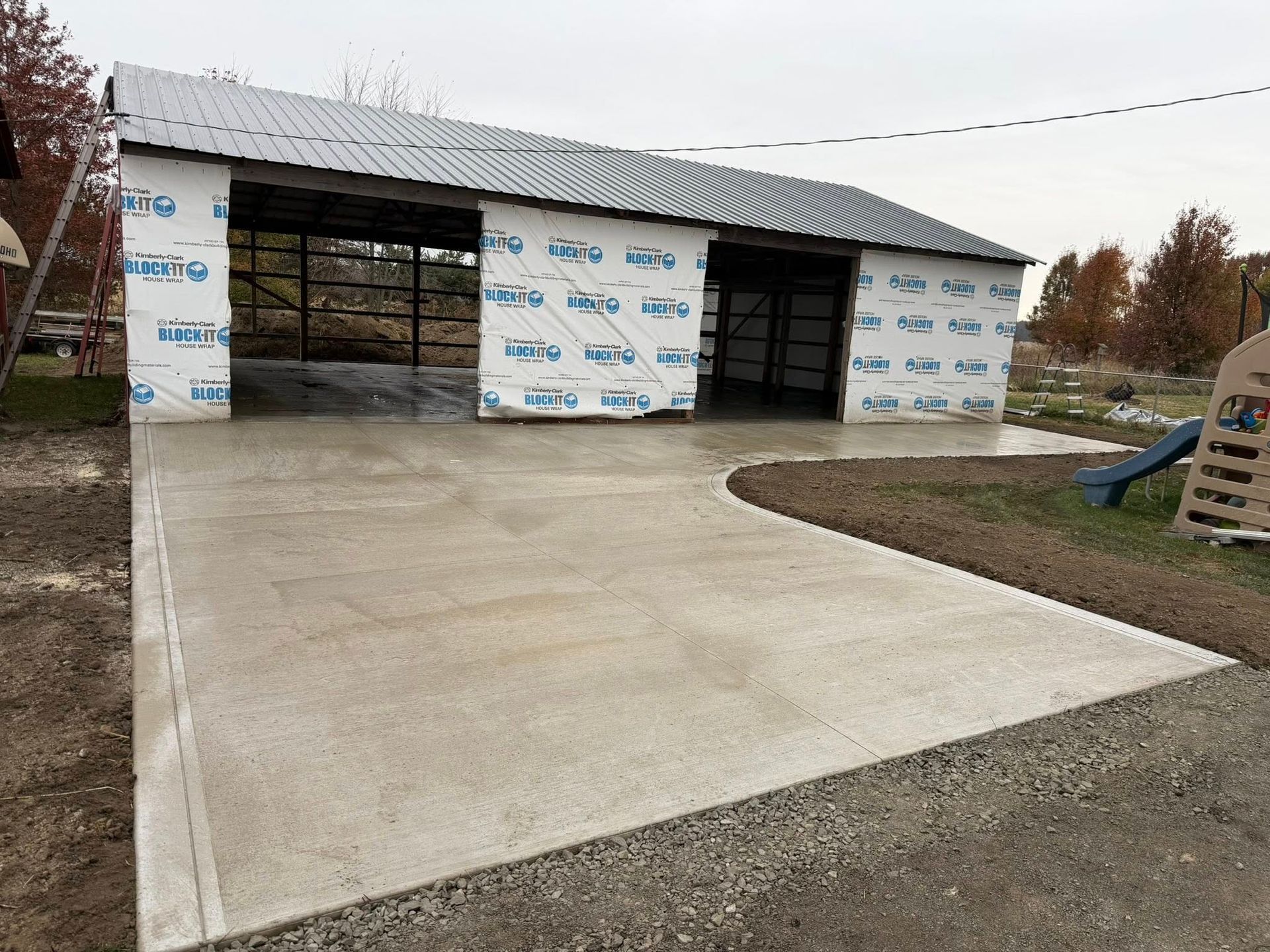 A newly poured concrete driveway leads to an unfinished, open-bay pole barn with white house wrap and a metal roof.