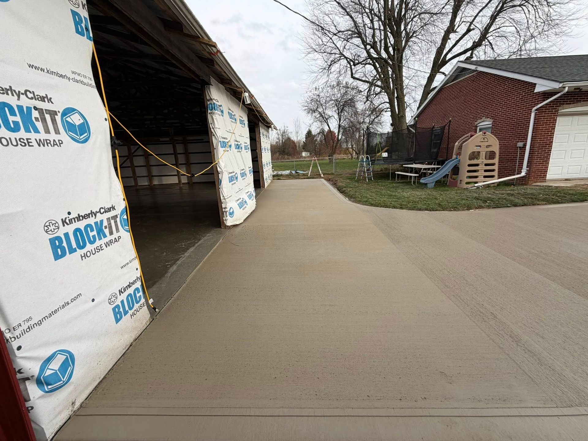 A new concrete walkway leads between a building under construction and a residential yard with a playground.