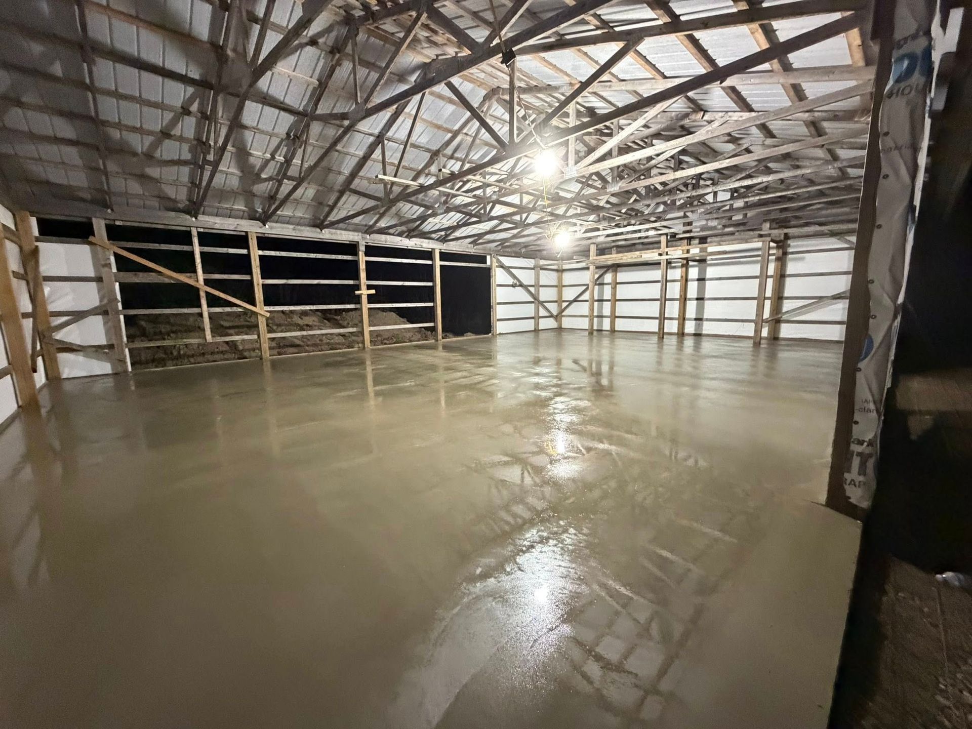 A freshly poured concrete floor inside a large, open-frame metal building with overhead wooden rafters.
