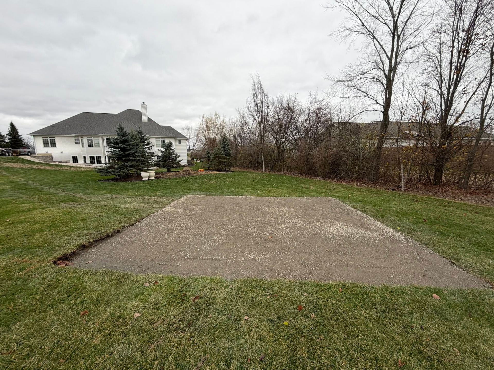 A rectangular patch of dirt and gravel sits in a grassy backyard, with a white house in the background.