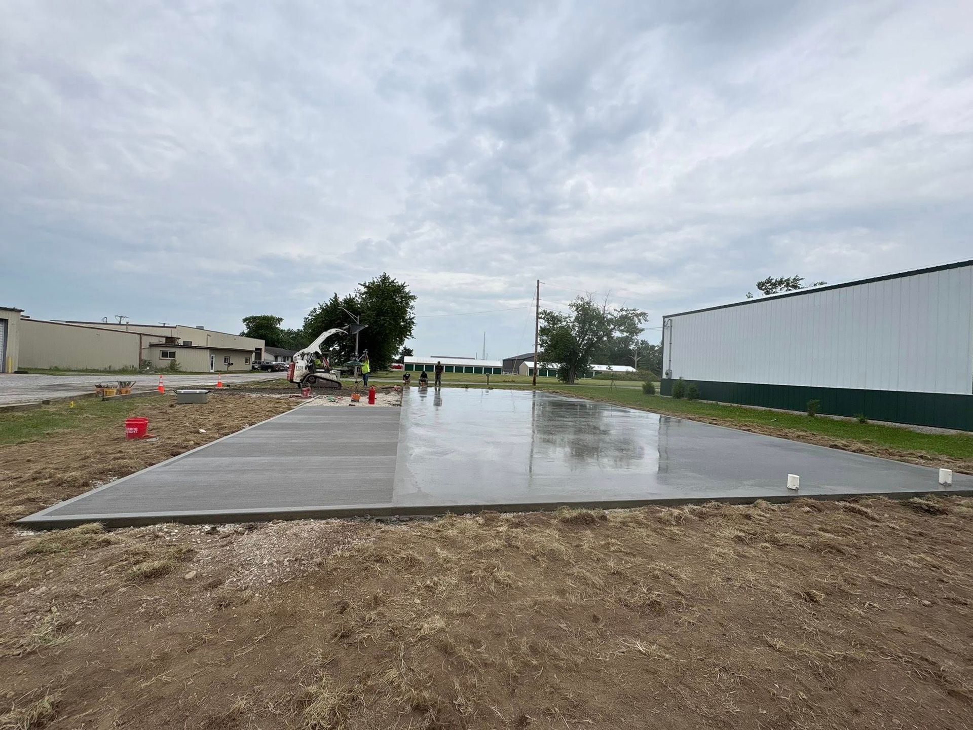 A freshly poured concrete slab sits in a dirt lot next to a white metal building under a cloudy sky.