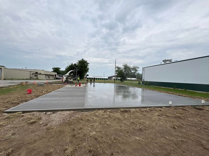 A newly poured concrete slab at a construction site with a white metal building nearby under a cloudy sky.